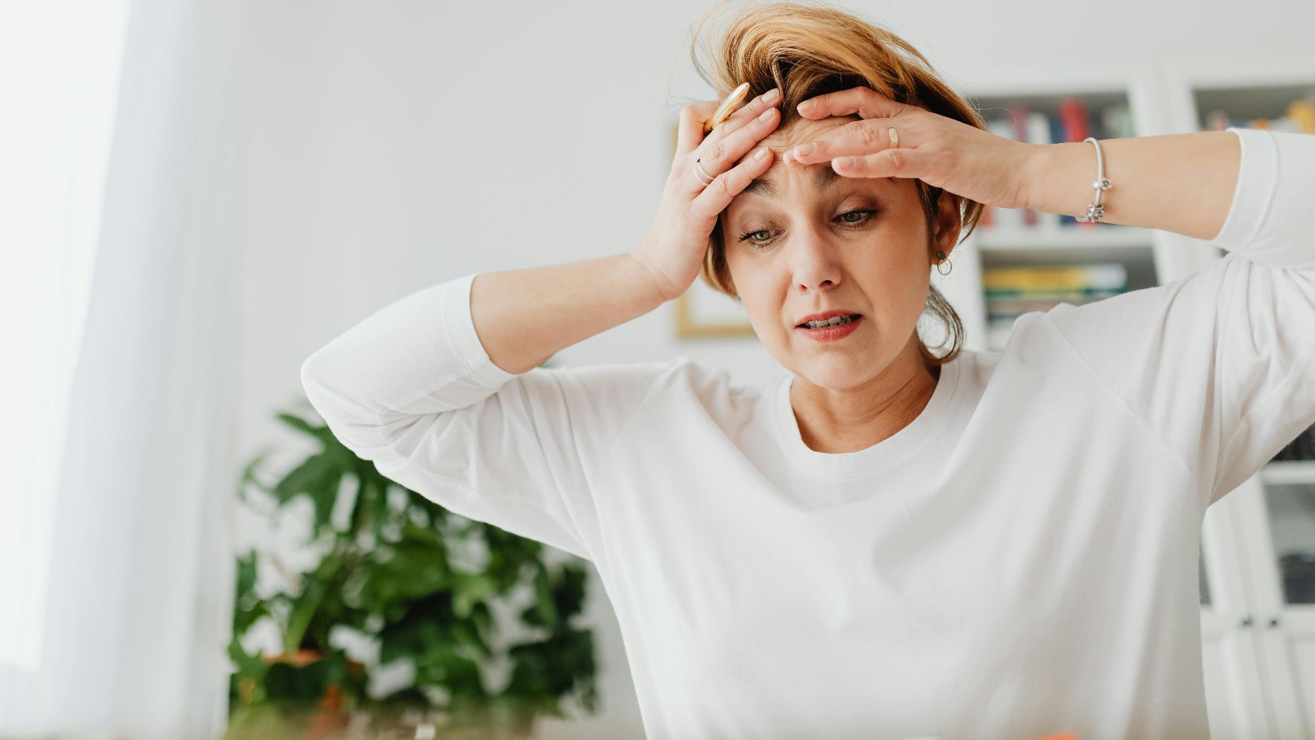 A worried woman indoors with hands on forehead, expressing stress.