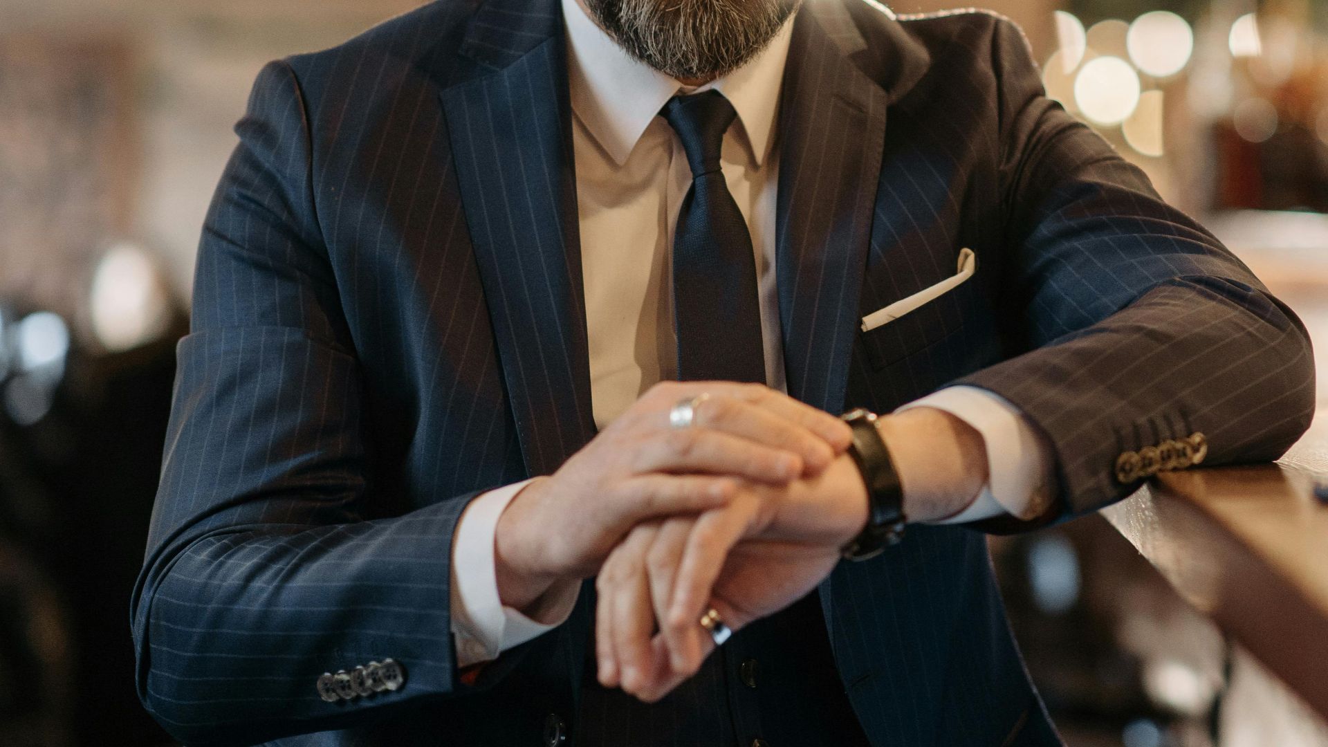 Stylish businessman in corporate attire enjoying whiskey at a bar, checking time.