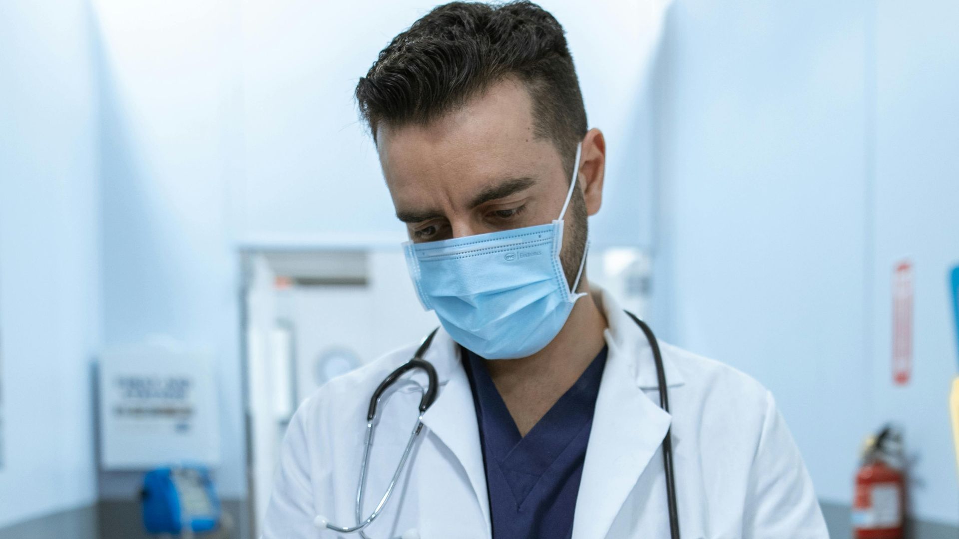 Medical professional wearing mask, writing on chart in hospital hallway.