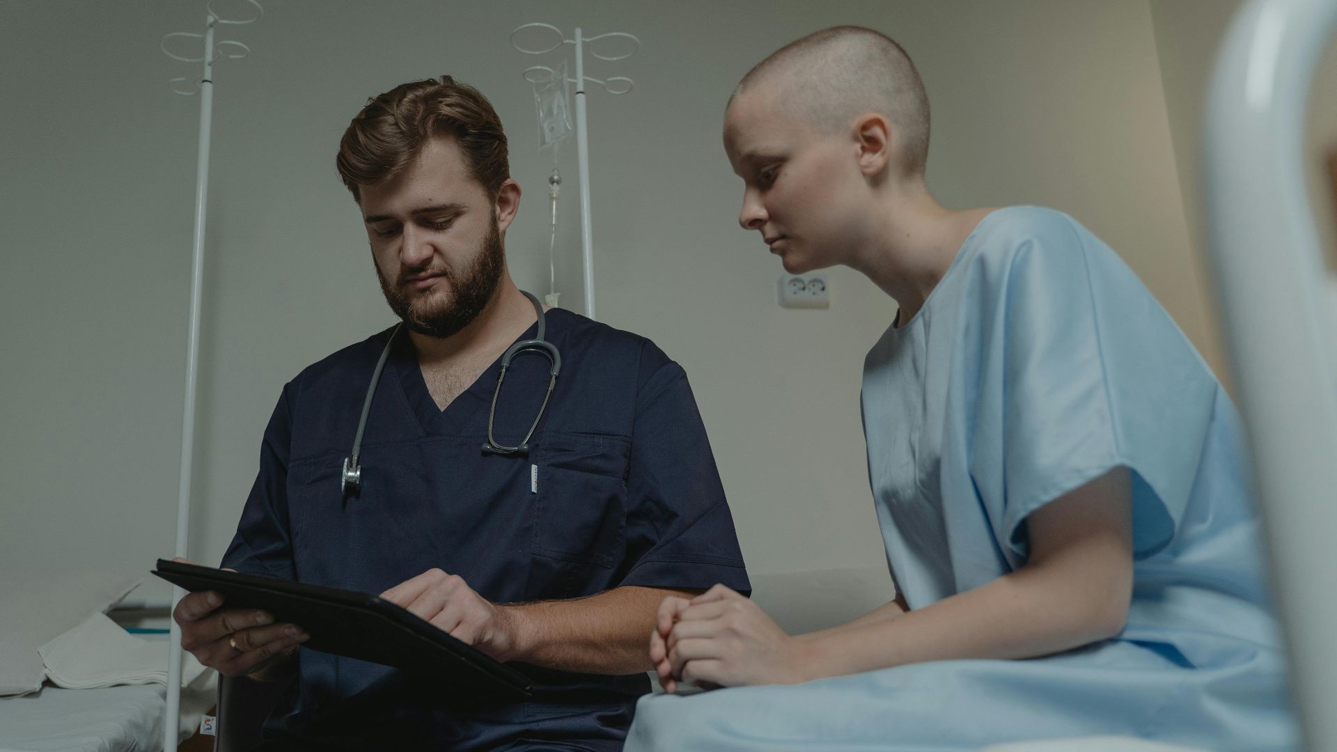 Doctor consulting with a young patient in a hospital room using a digital tablet.