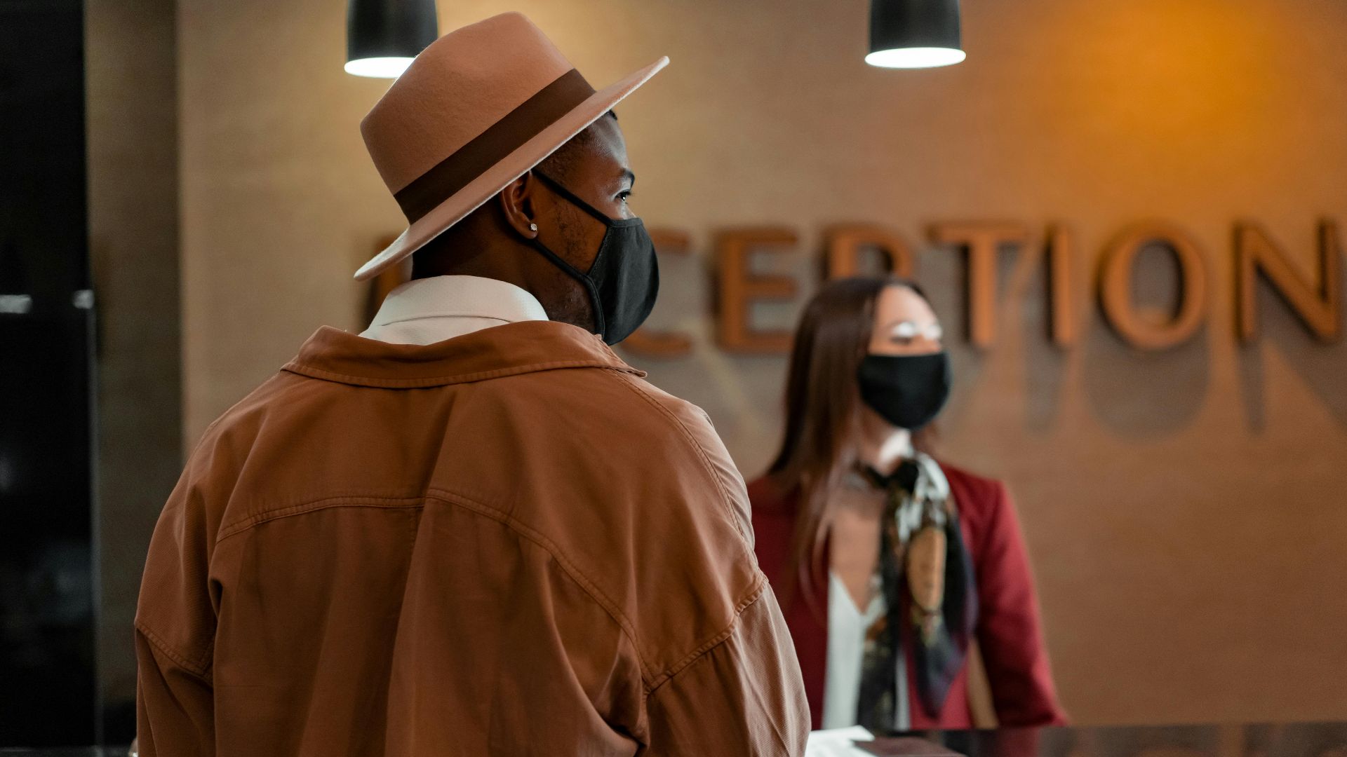 Man in mask at hotel reception during pandemic, interacting with masked receptionist.