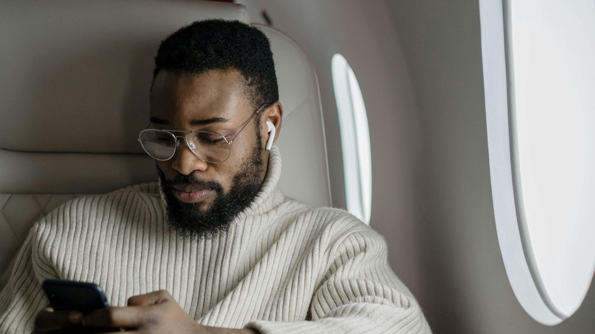 Black businessman texting on smartphone in a private jet's window seat.