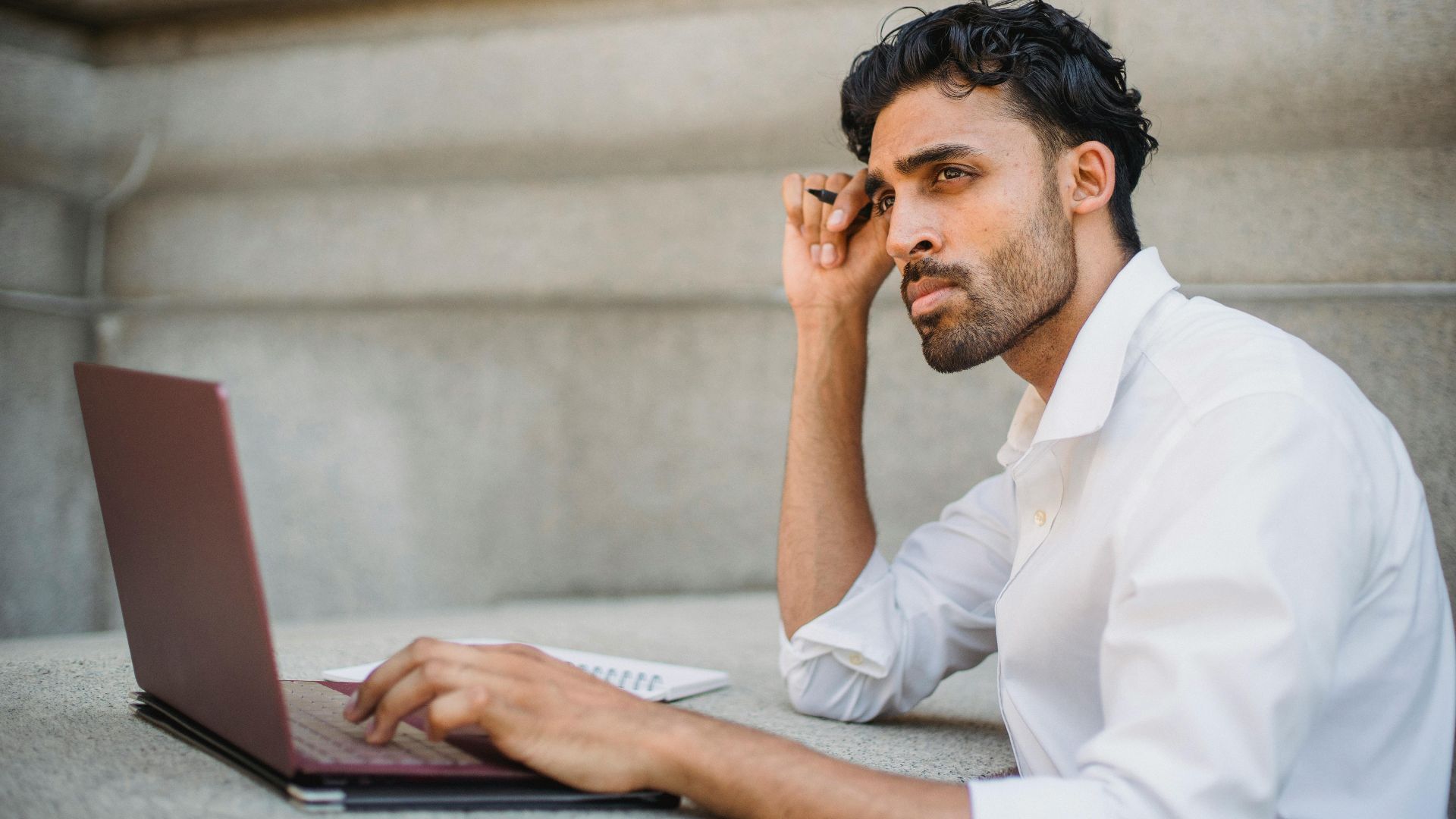 Professional man in a white shirt working on a laptop, contemplating ideas in an urban setting.