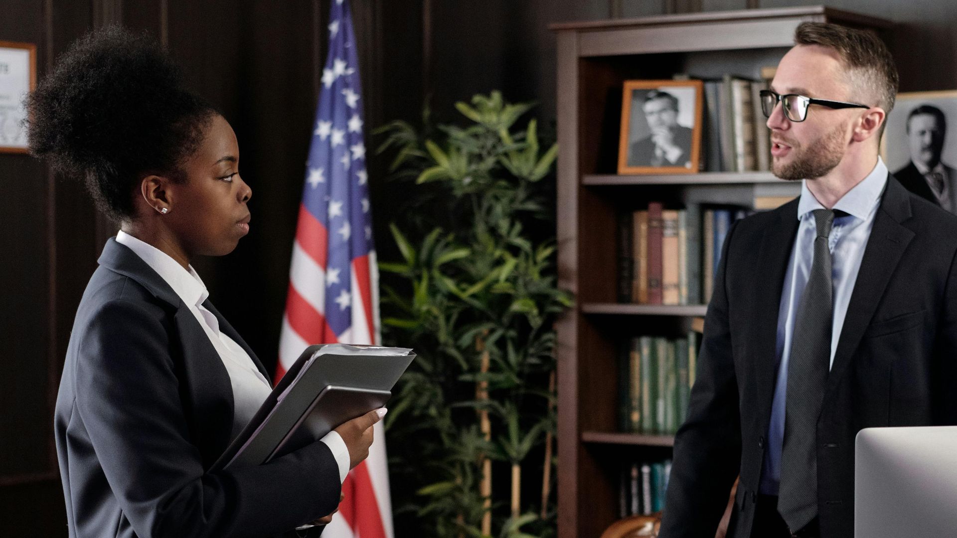 Two colleagues in a formal office setting engaged in a discussion with American flag in background.