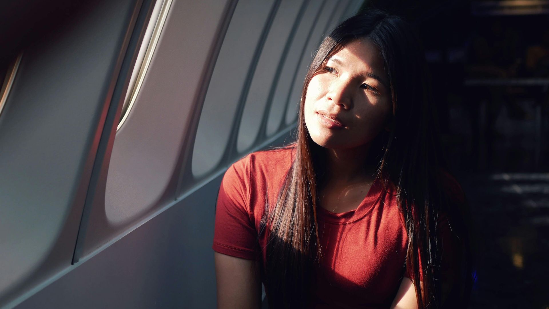 A woman in a red shirt sits in an airplane, gazing thoughtfully out the window as sunlight illuminates her face.