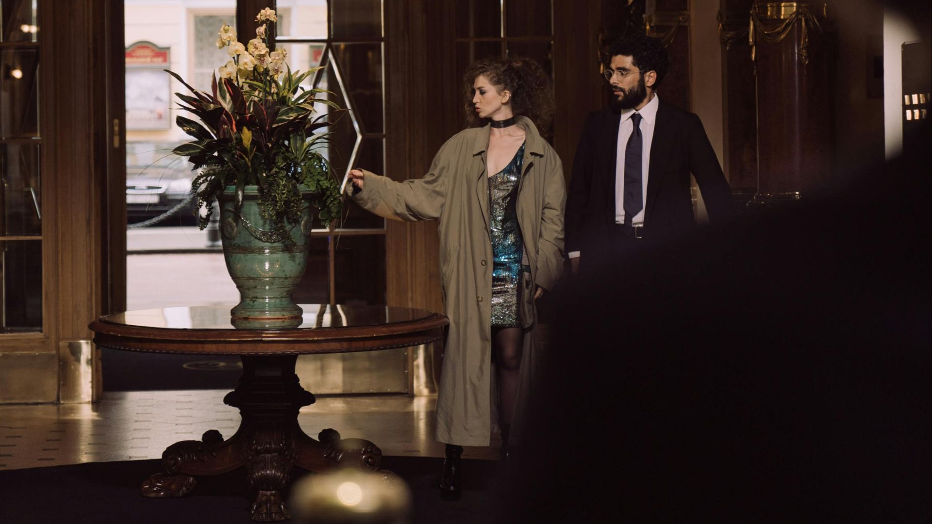 Luxurious hotel lobby featuring an ornate chandelier and an elegant couple admiring a flower arrangement.