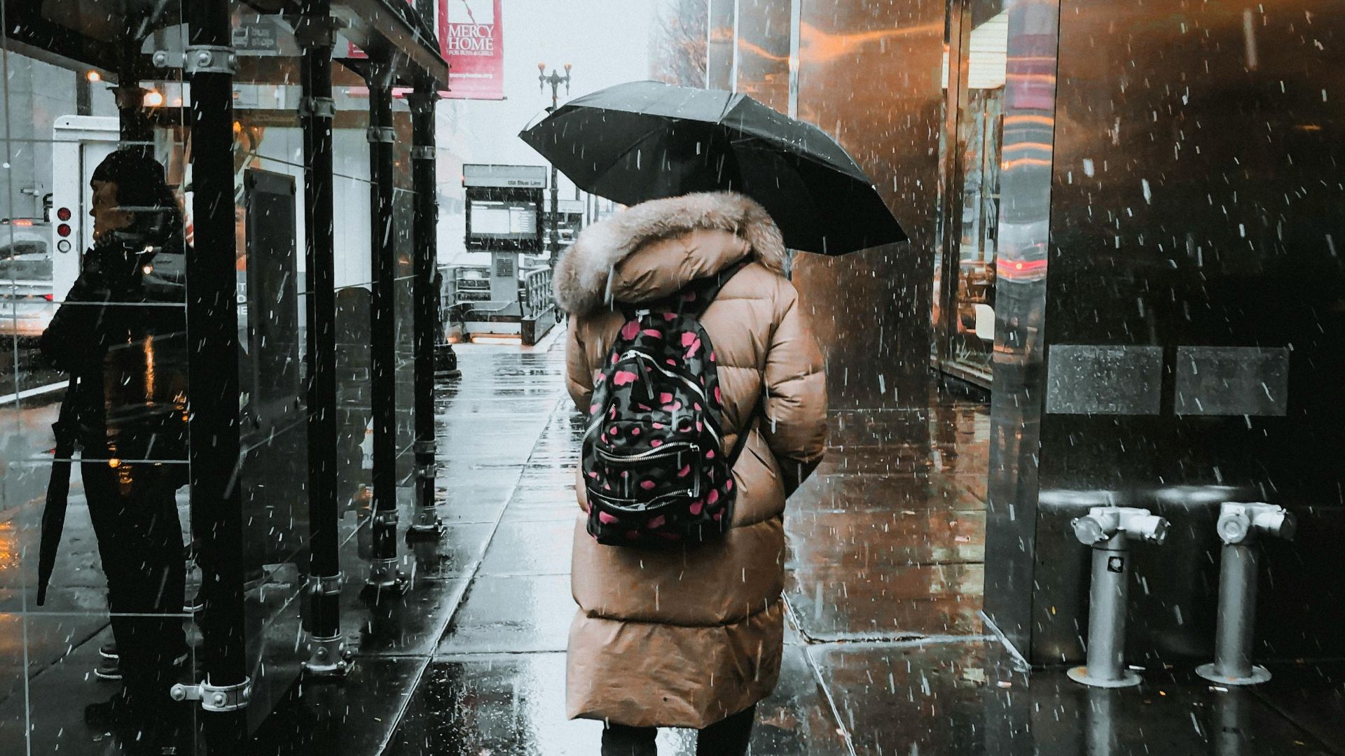 A woman walks on a rainy city street, carrying an umbrella and wearing a winter coat. Reflections on wet pavement add depth.