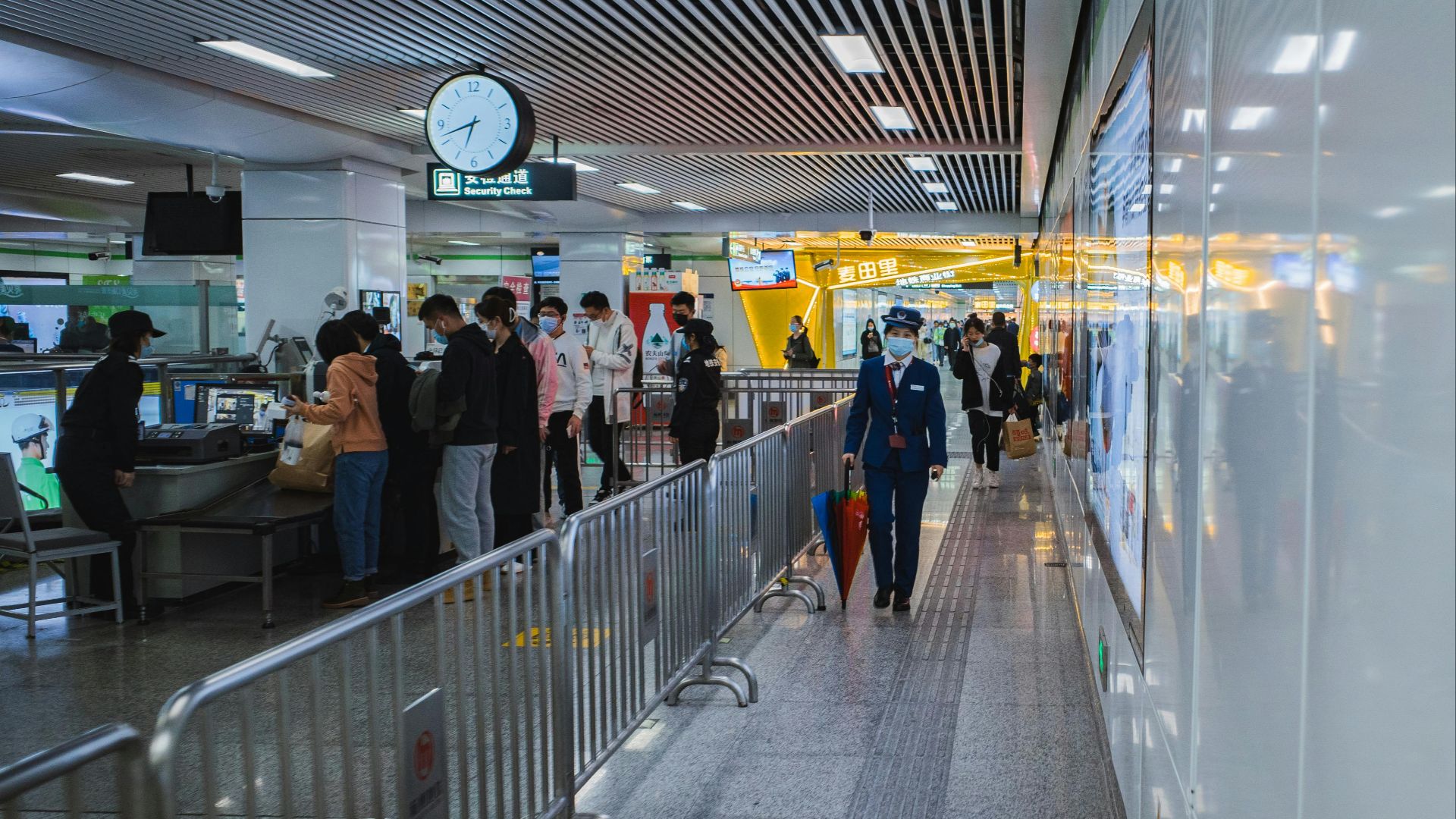 Travelers passing through airport security with staff and metal railings visible.