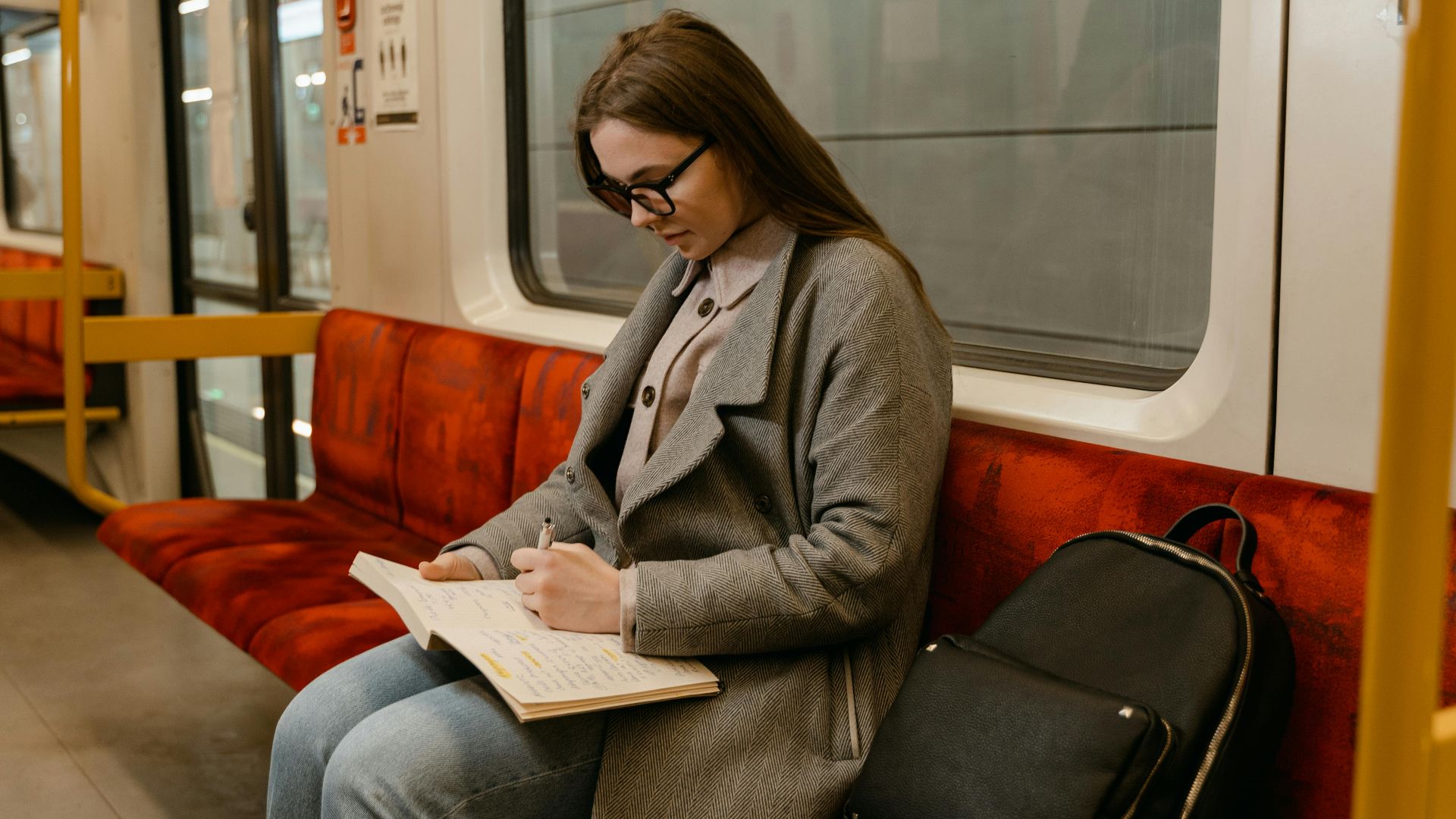 A woman in glasses and a gray coat writes in a notebook while commuting on a train.