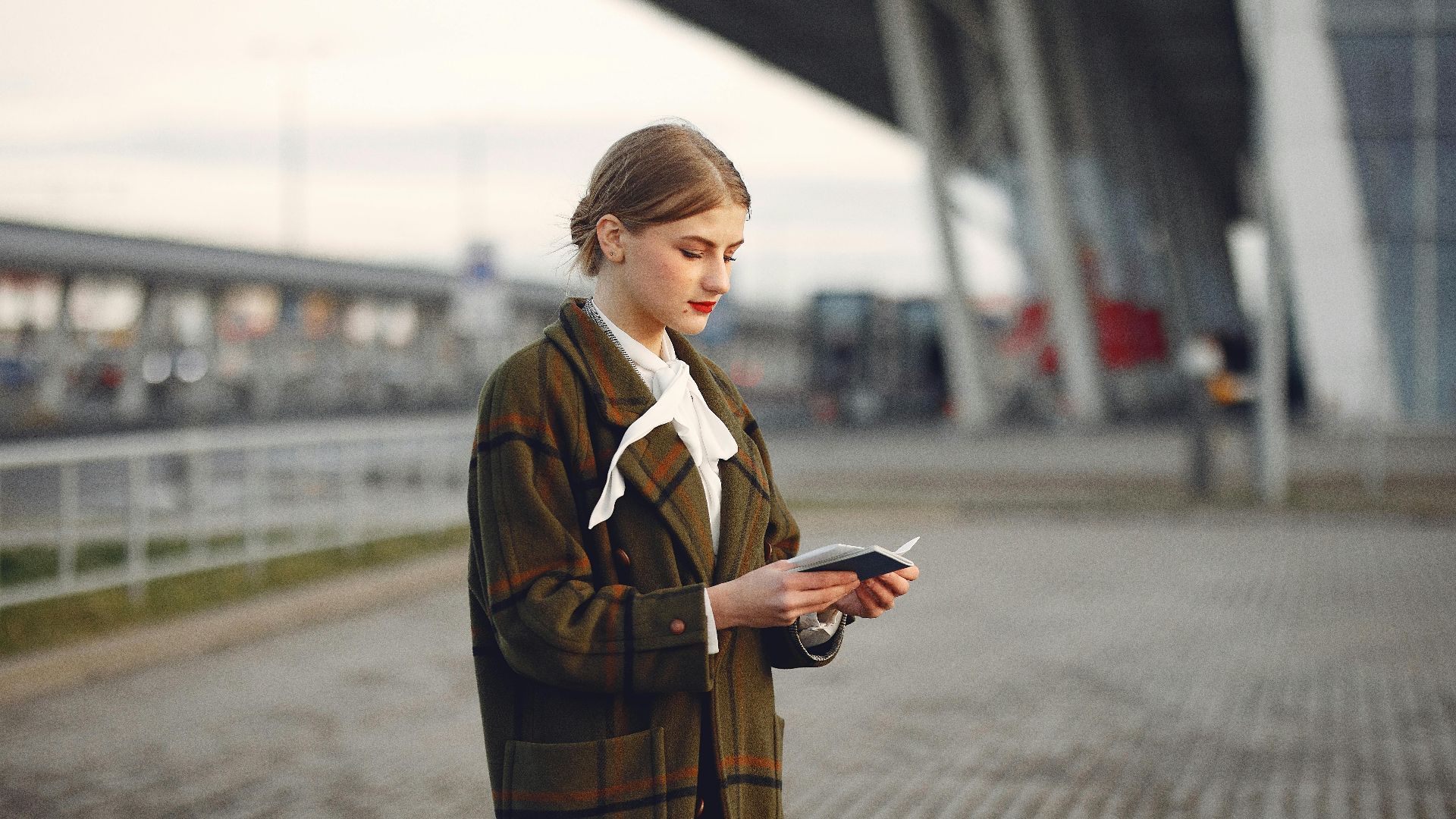 Attentive female passenger wearing trendy plaid coat and white blouse checking passport and ticket standing on pavement near modern building of airport outside
