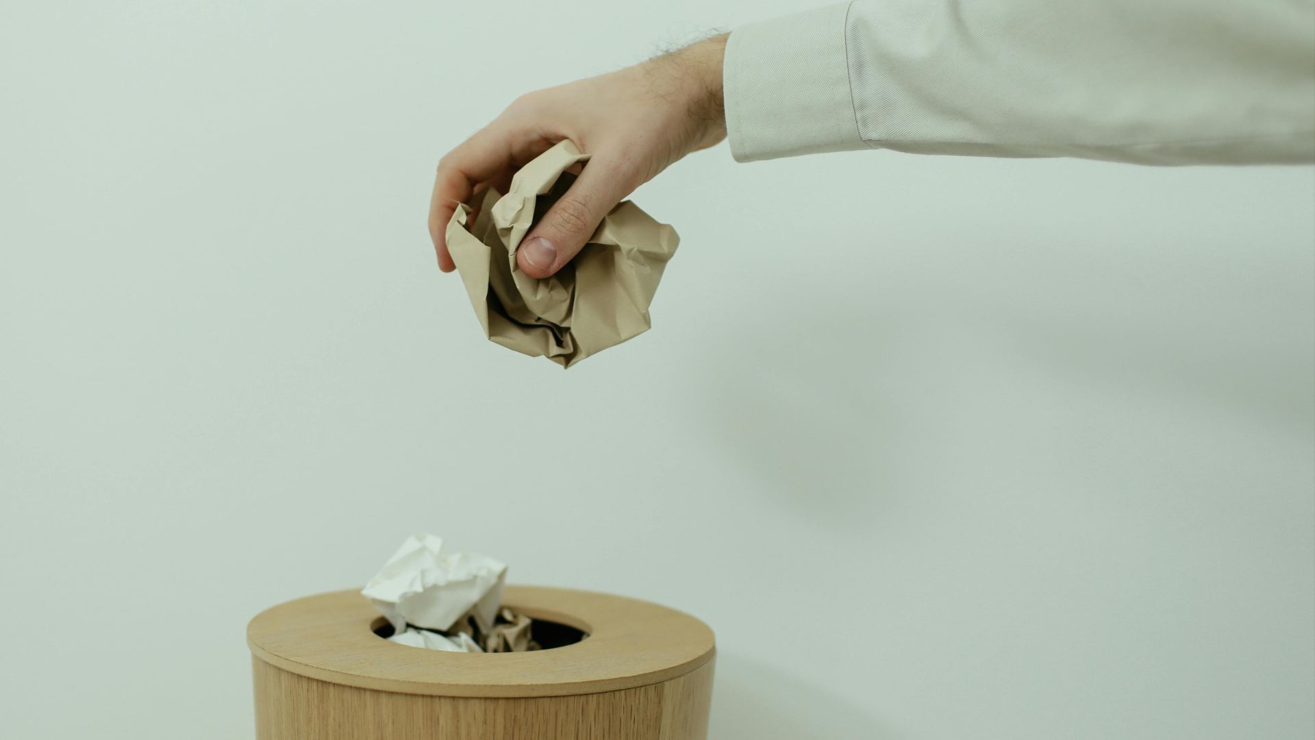 A person's hand disposes of crumpled paper into a wooden trash bin indoors.
