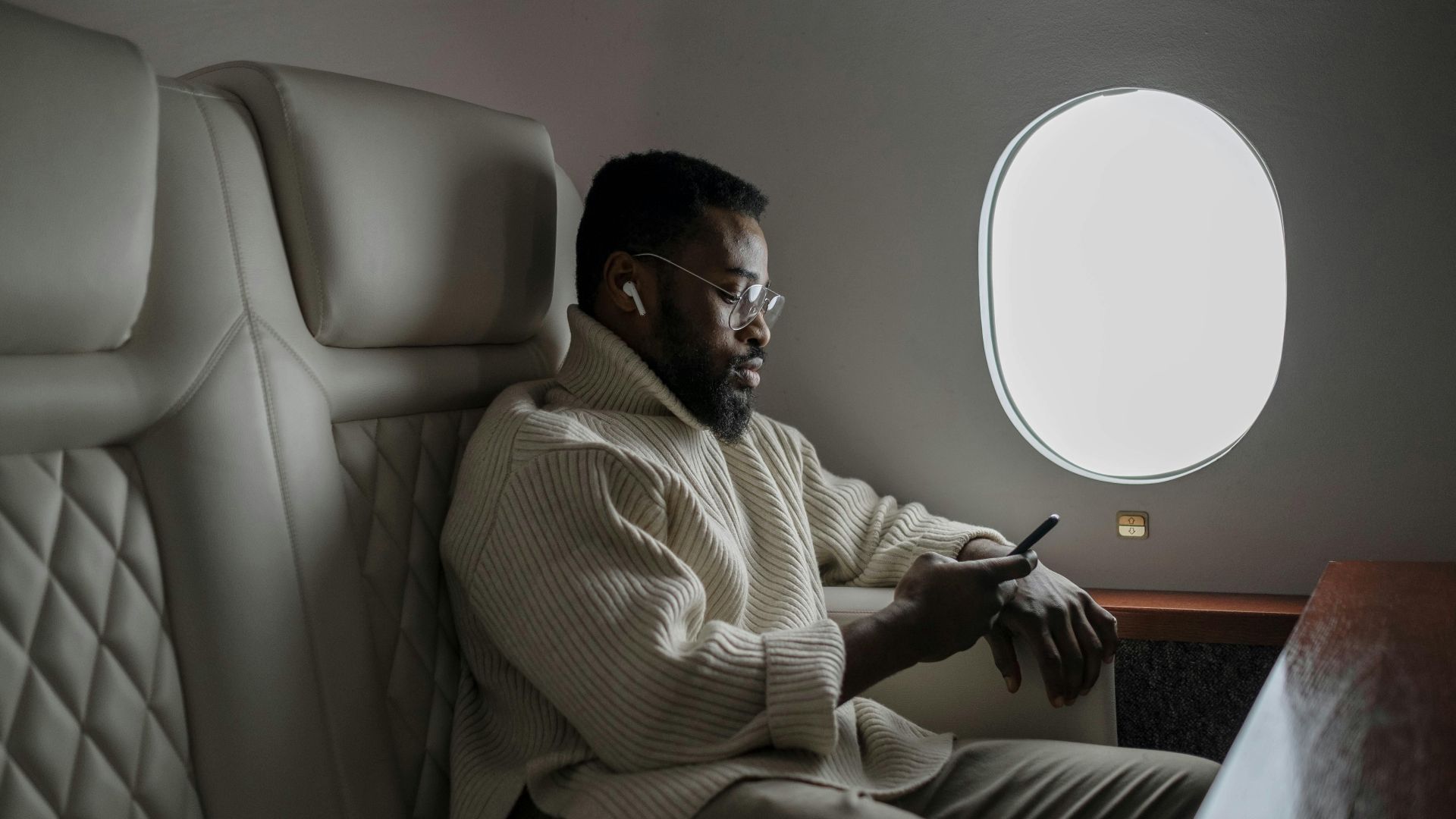 Bearded man in cozy sweater using smartphone on airplane, enjoying in-flight entertainment.