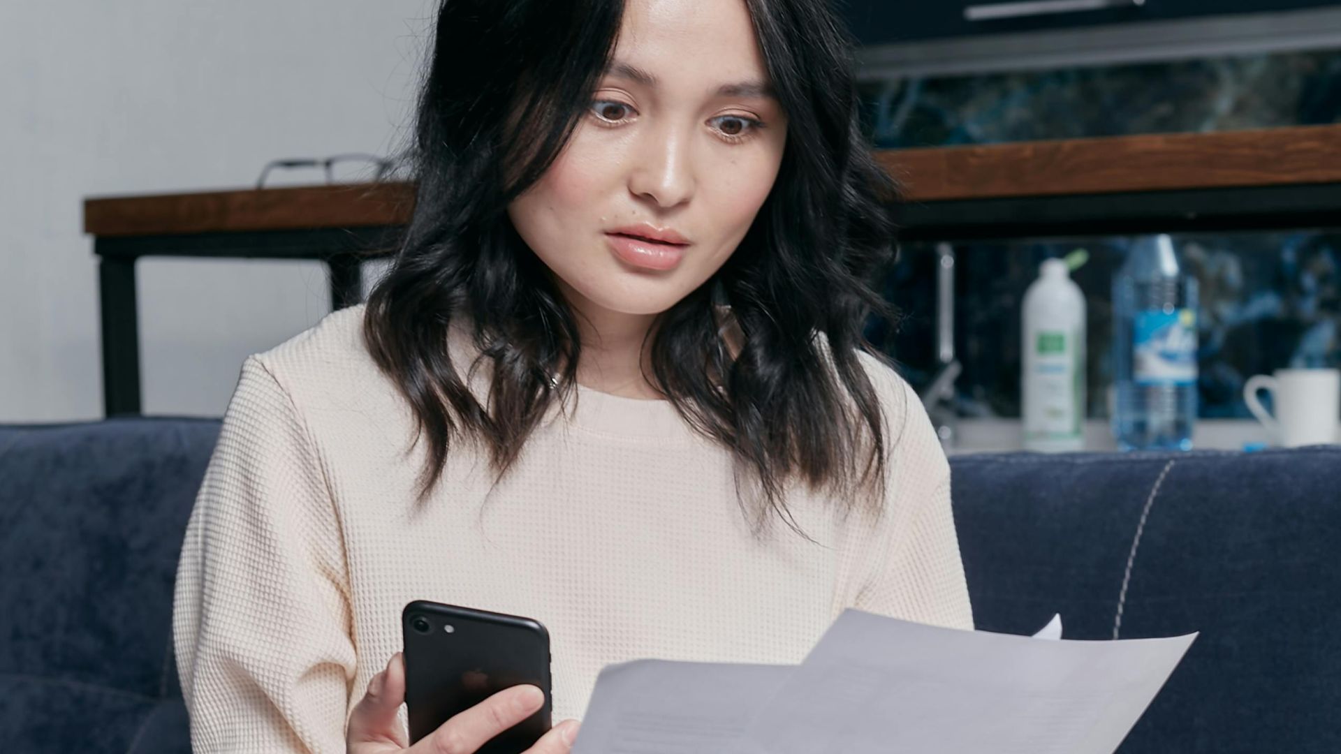 Young woman reviews documents and phone indoors, showcasing concern while seated on a couch with scattered papers. Business concept.