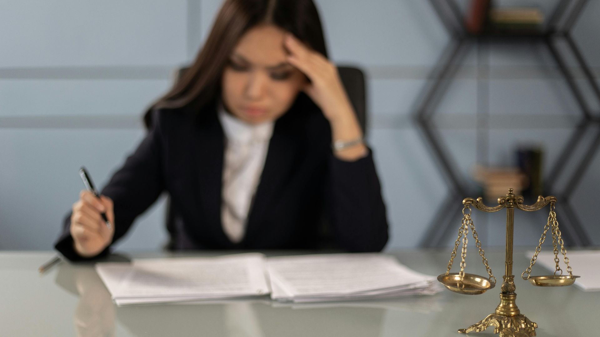 A fatigued female lawyer reviewing documents with a justice scale nearby, emphasizing stress in legal work.