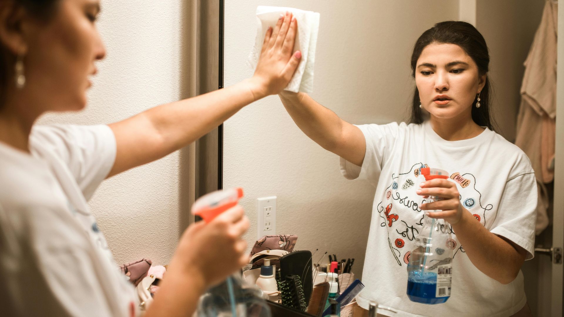 An adult woman cleaning a bathroom mirror with a spray bottle and cloth, focused on hygiene.