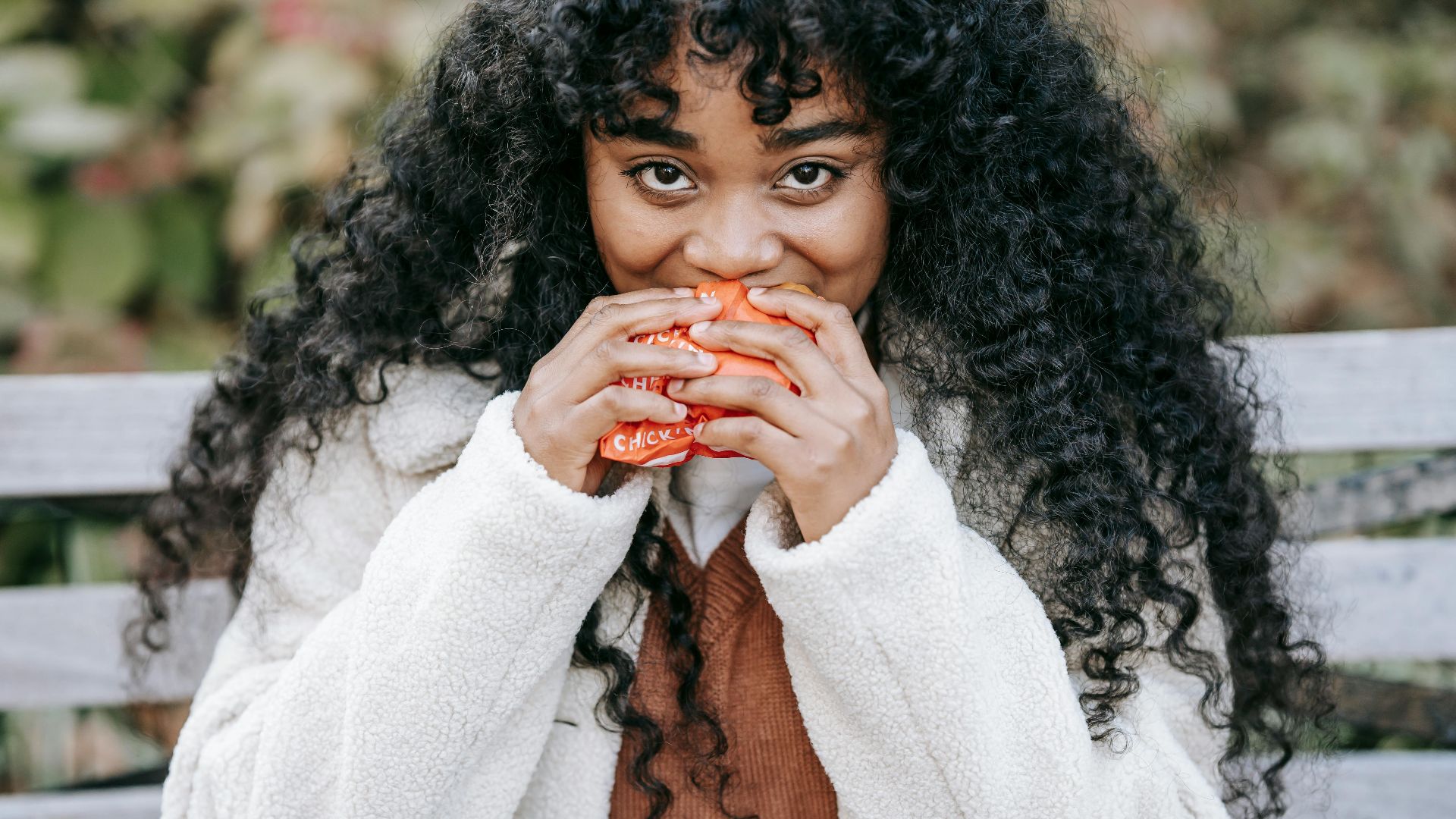 Crop hungry African American female in warm clothes biting delicious fresh burger and looking at camera while sitting on bench in park