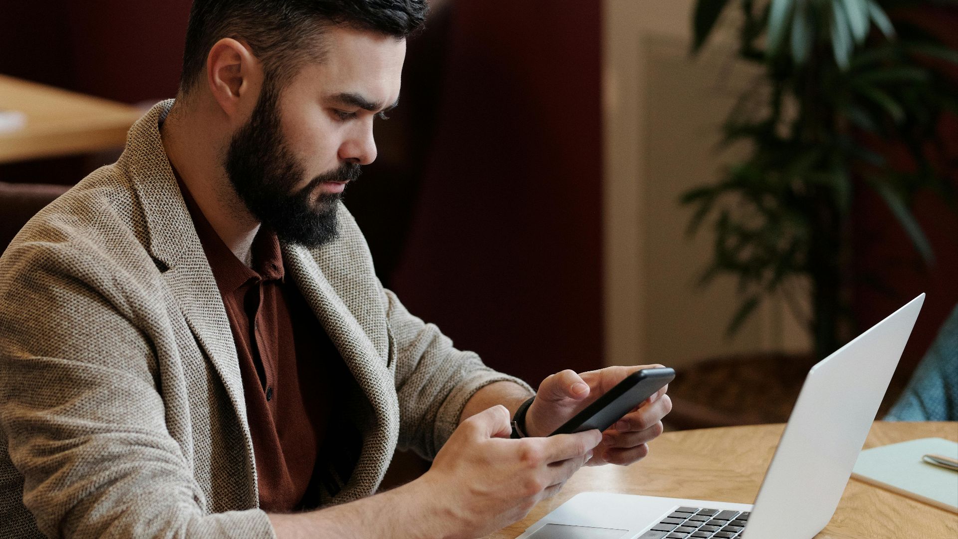 Bearded man using smartphone and laptop at café with coffee, focused on remote work.