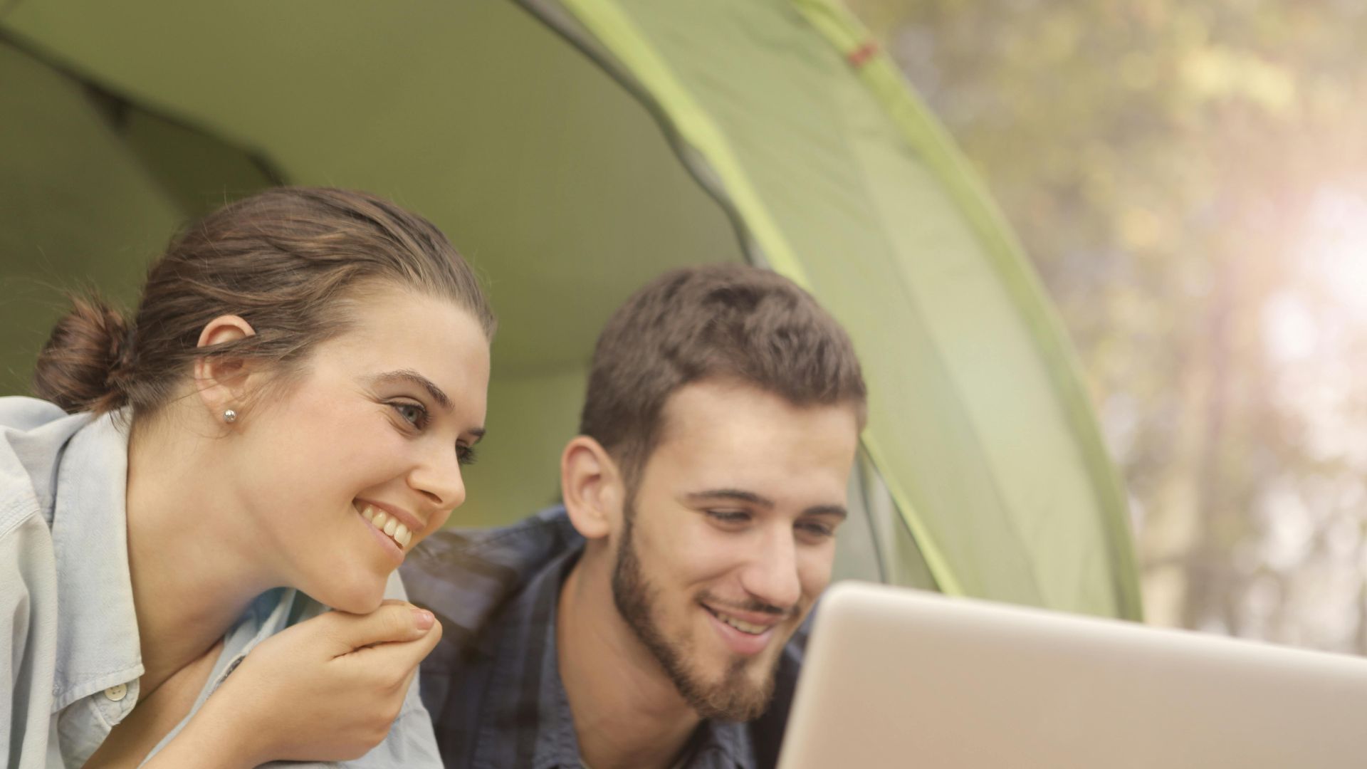 A happy couple in a tent enjoying leisure time with a laptop in a sunny outdoor setting.