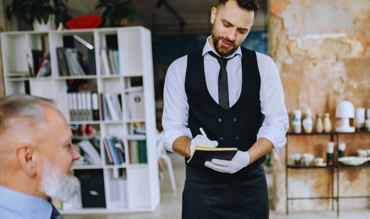 Waiter Taking Order from Customers at a Glass Table in an Elegant Interior with Textured Walls