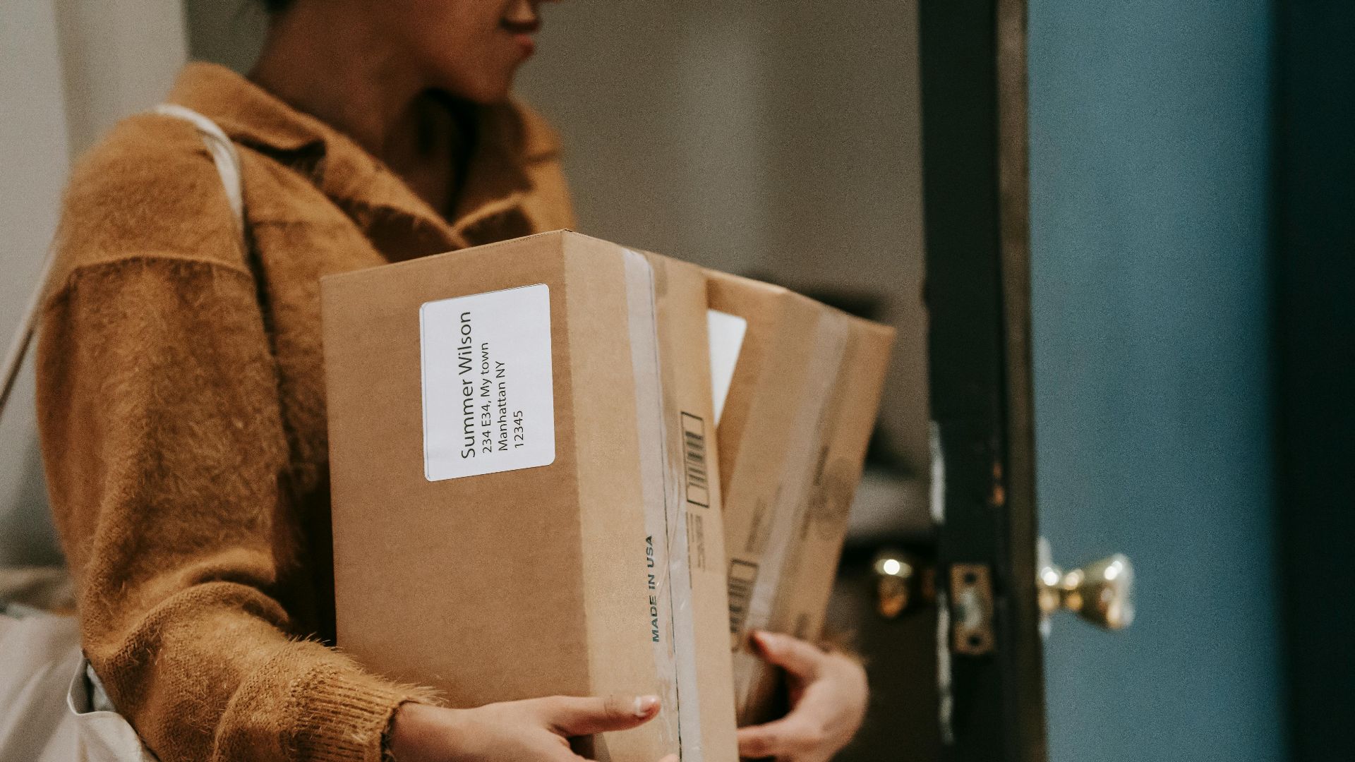 Crop ethnic female walking into open door of apartment with carton boxes with goods from delivery