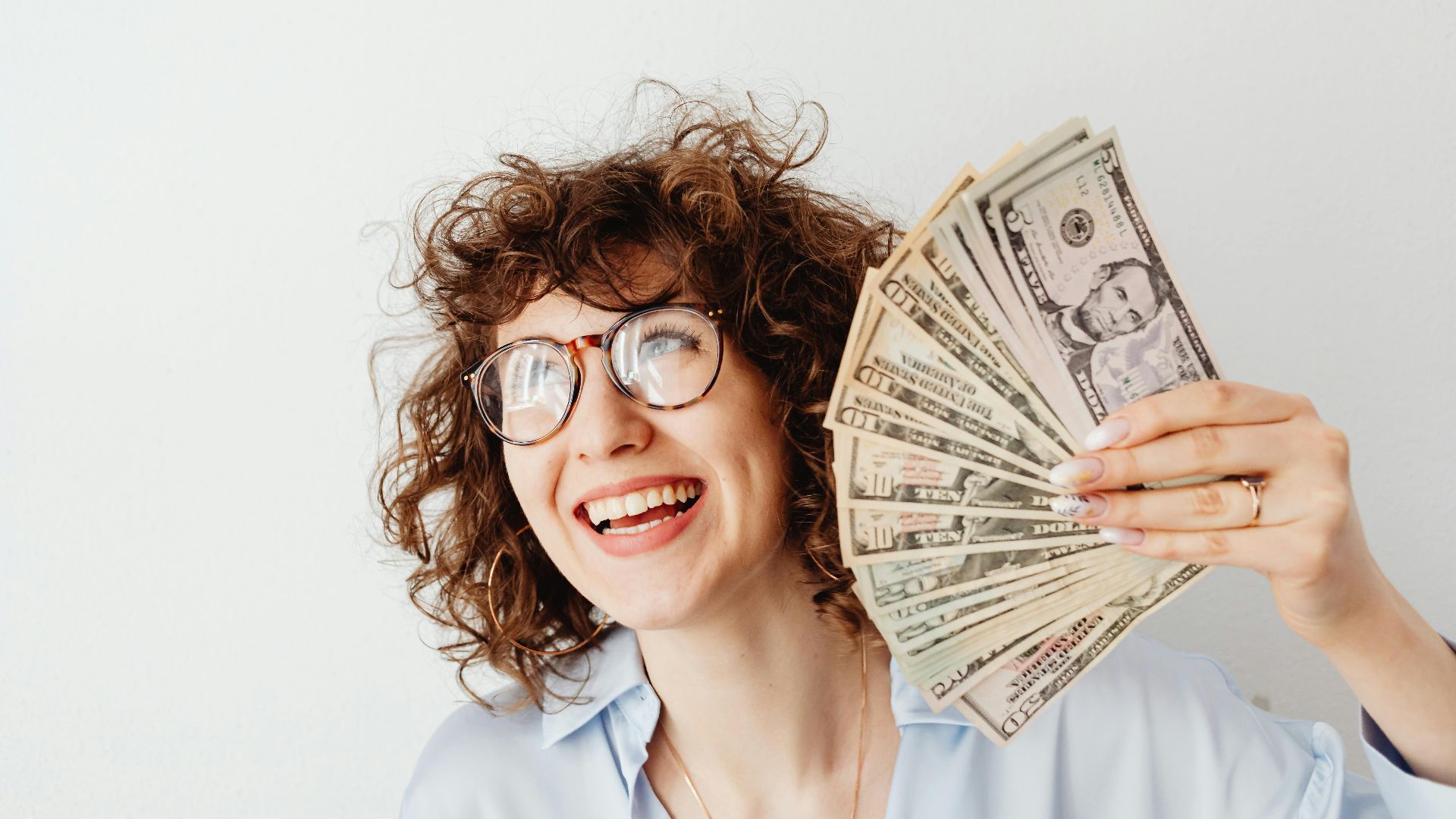 Happy woman with curly hair and glasses holding US dollar bills against a white background.