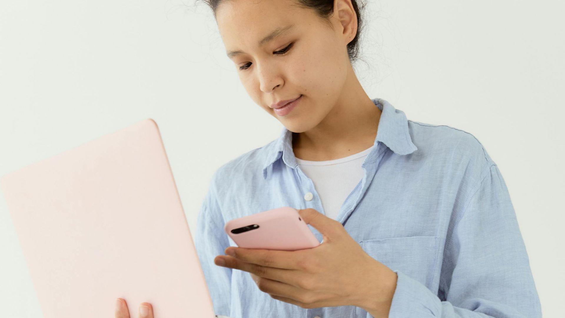 Asian woman in blue shirt multitasking with a laptop and smartphone indoors.