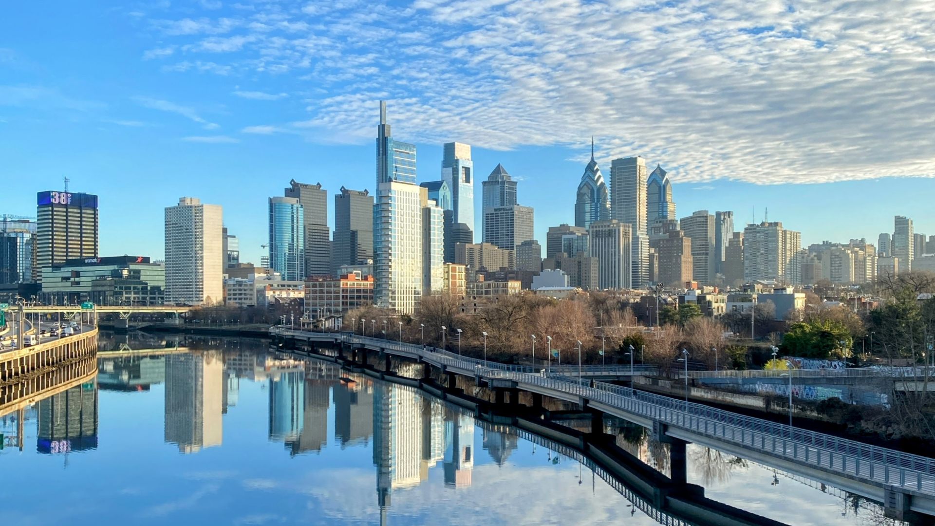 This photo shows a skyline view of Philadelphia, Pennsylvania, as seen from the South Street Bridge on December 31, 2024. The Schuylkill River and Schuylkill Banks Boardwalk are visible in the foreground, and a scattering of clouds can be seen reflecting in the water.