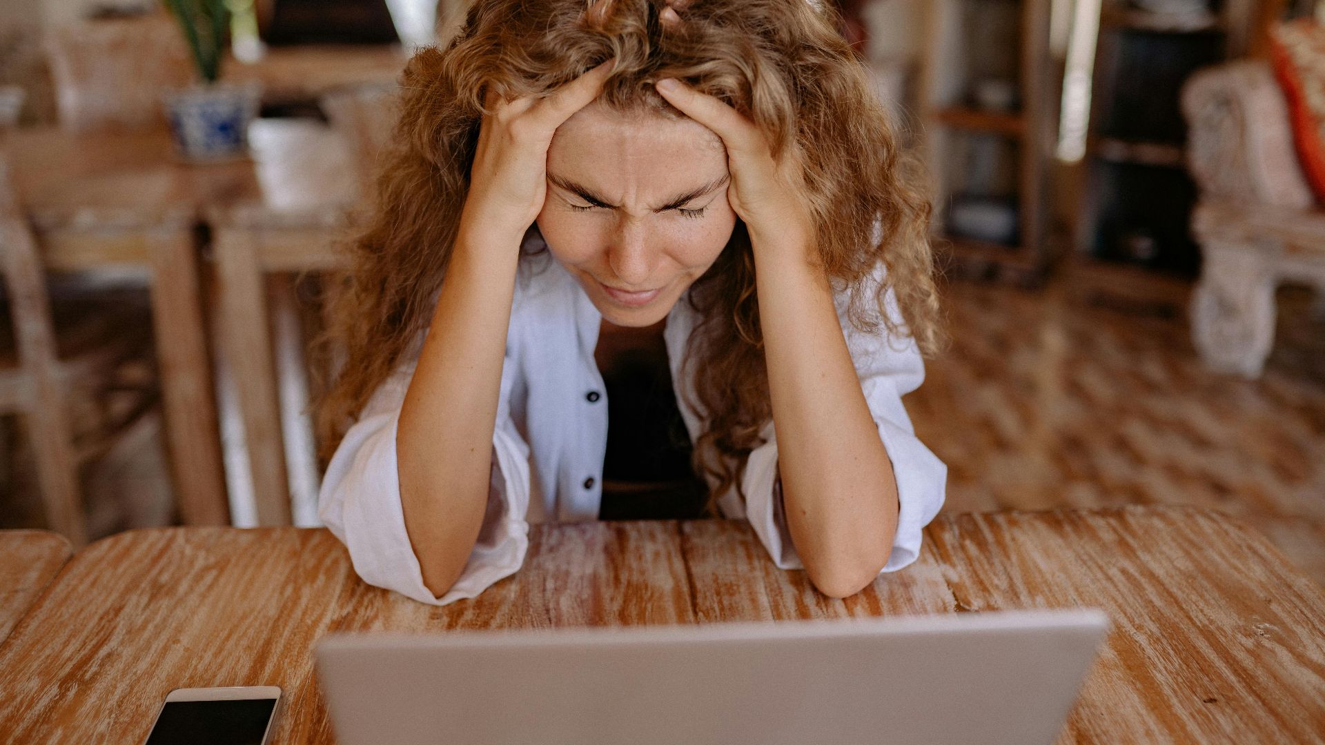 A stressed woman sits at a wooden table, frustrated with her laptop work.