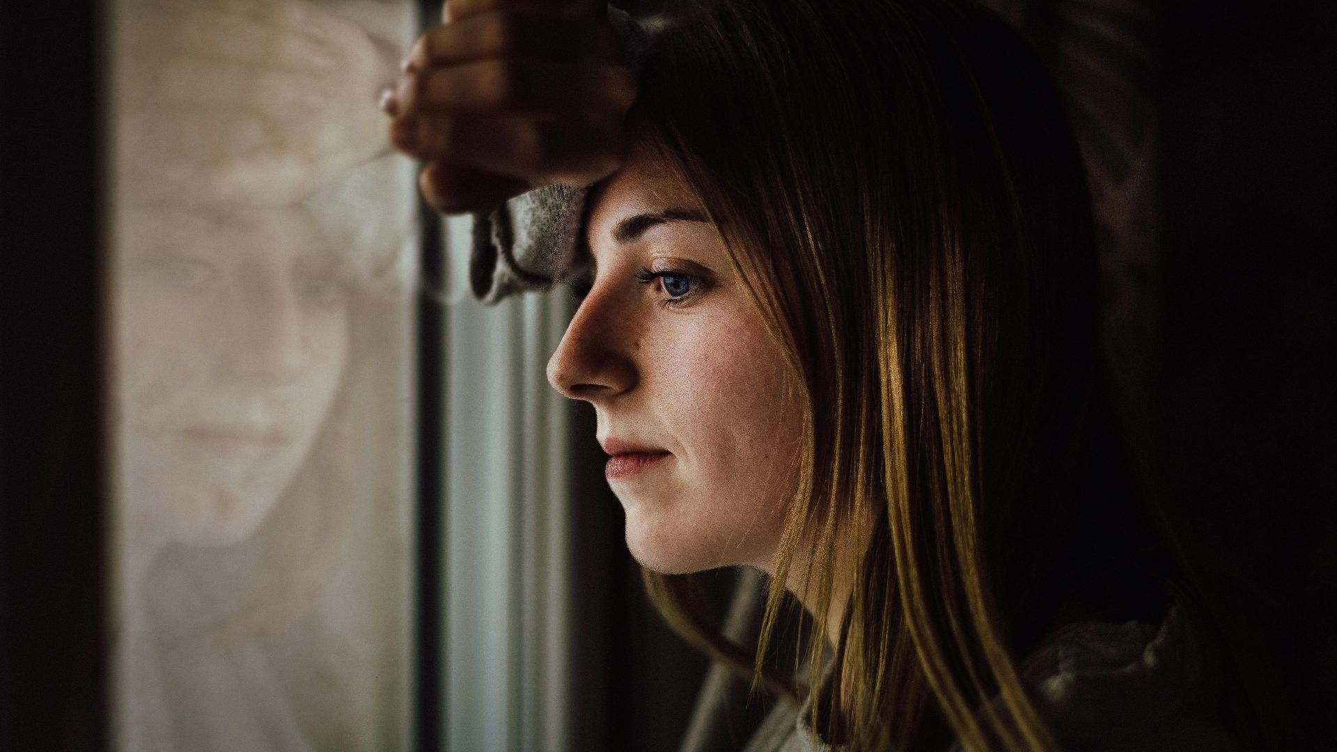 A contemplative young woman looks out a window with a reflective expression, indoors.