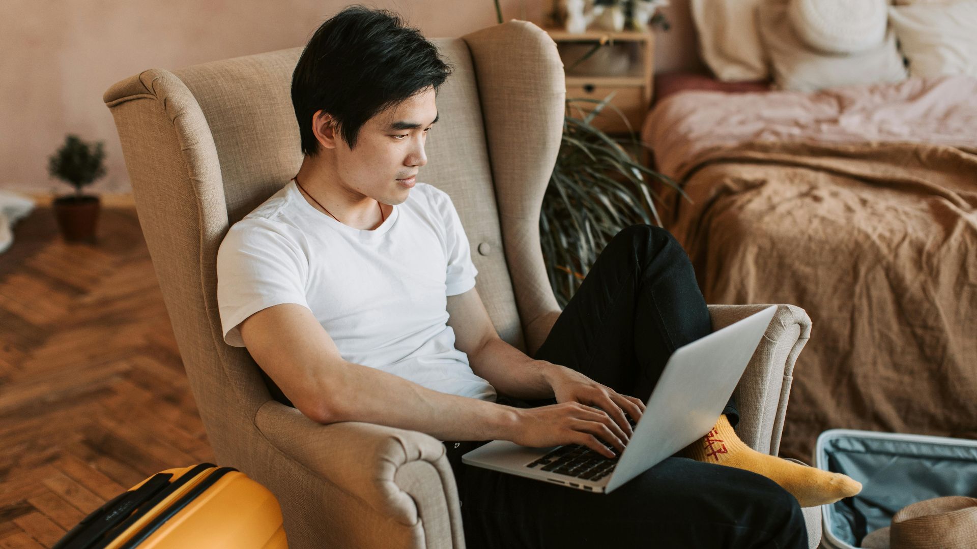 A man sitting comfortably in an armchair, using a laptop in a cozy home interior.