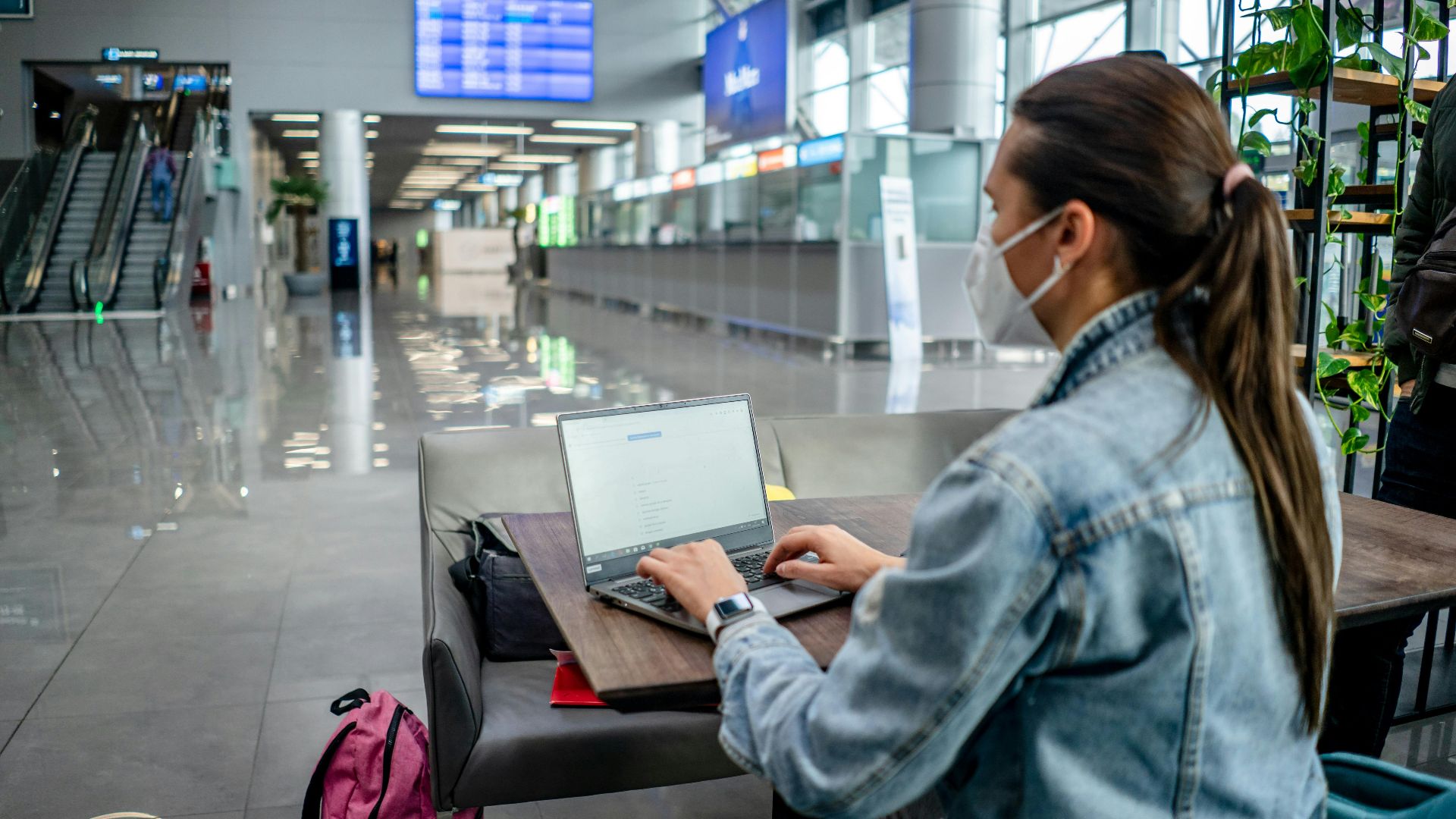 Side view of concentrated female freelancer in protective mask typing on netbook while sitting at table in modern airport terminal