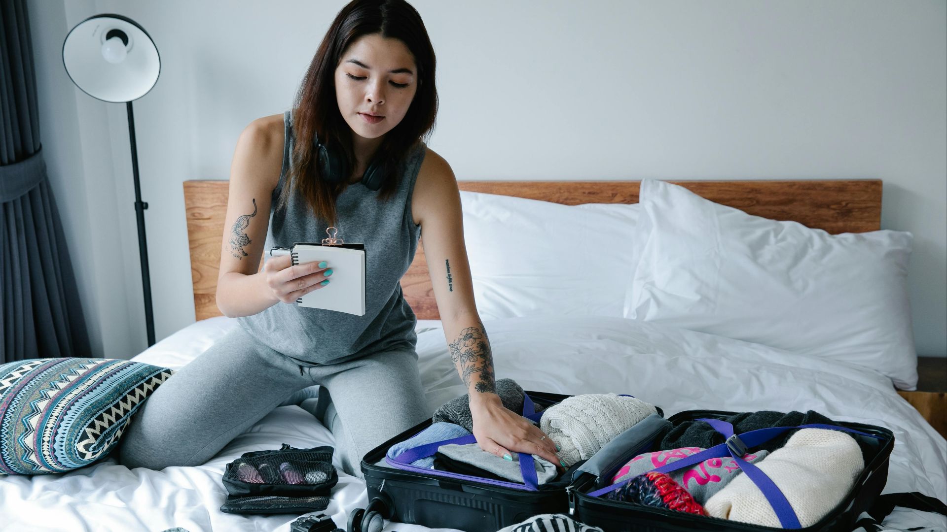 Woman sitting on bed packing a suitcase and taking notes for her travel plan.