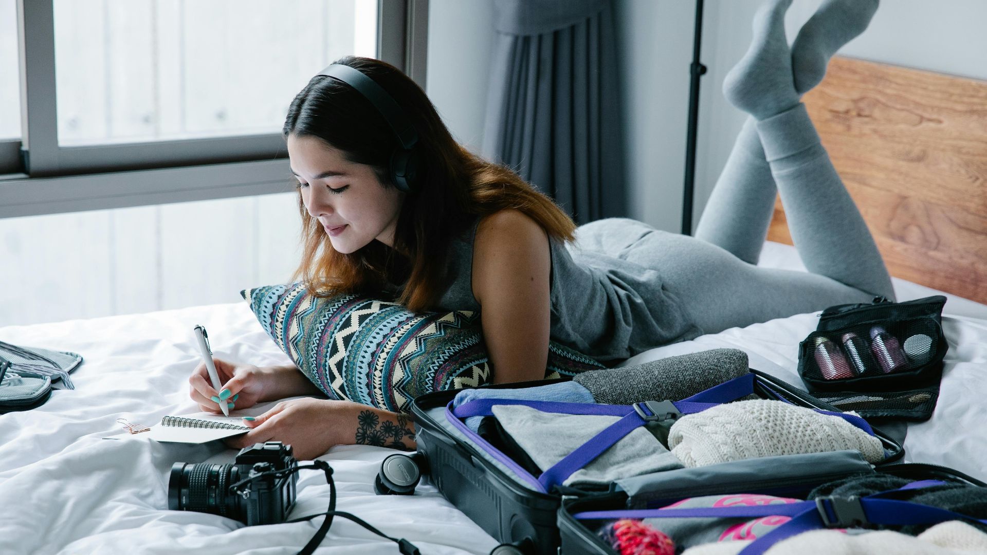 Woman lying on bed with headphones, writing in notebook, and packing suitcase.