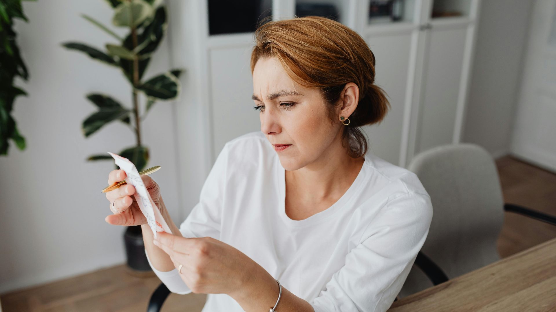 A concerned adult woman examines a receipt while sitting at her office desk, indicating financial scrutiny.