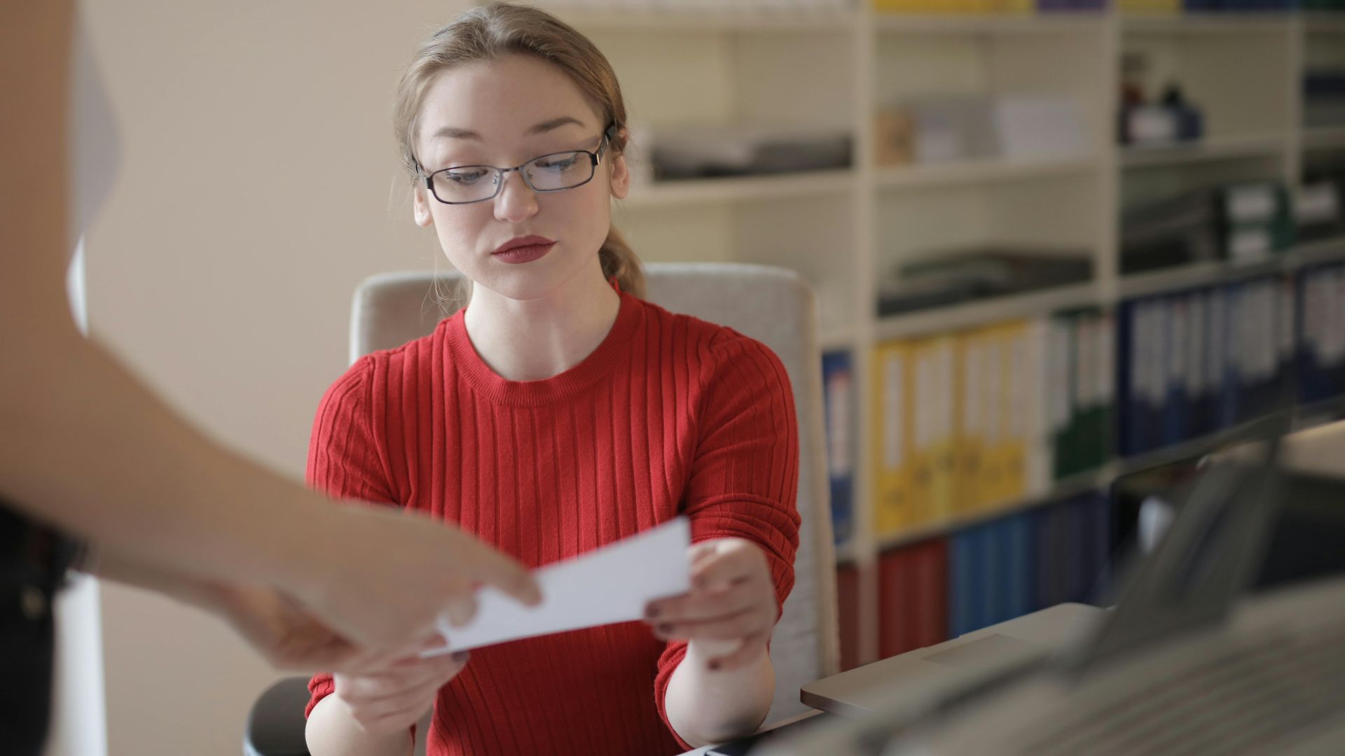 Serious young female employee in casual clothes and eyeglasses reading attentively document while working in office with colleague