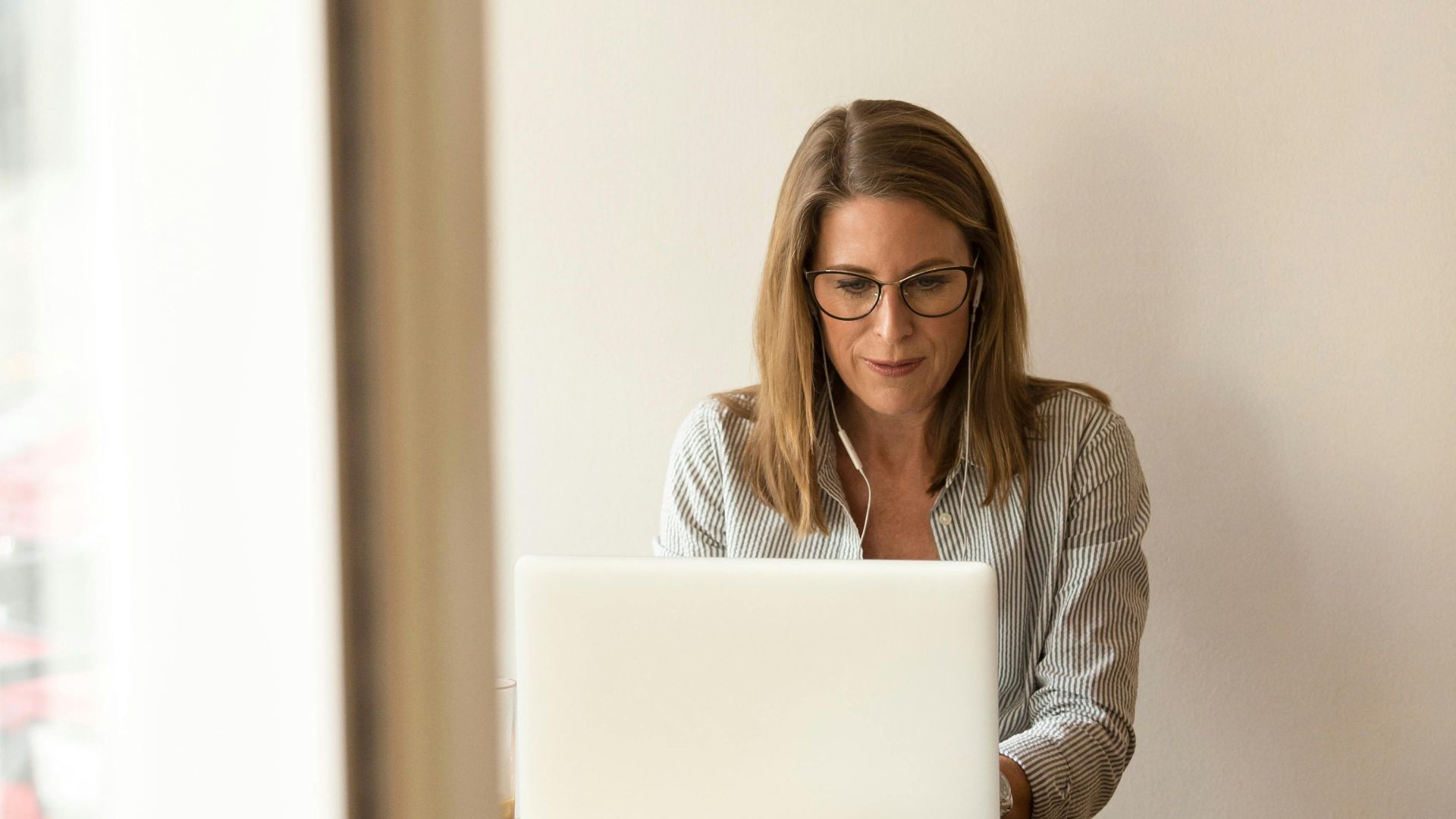 Businesswoman working remotely on a laptop at home, enjoying natural light in a modern, minimalist setting.