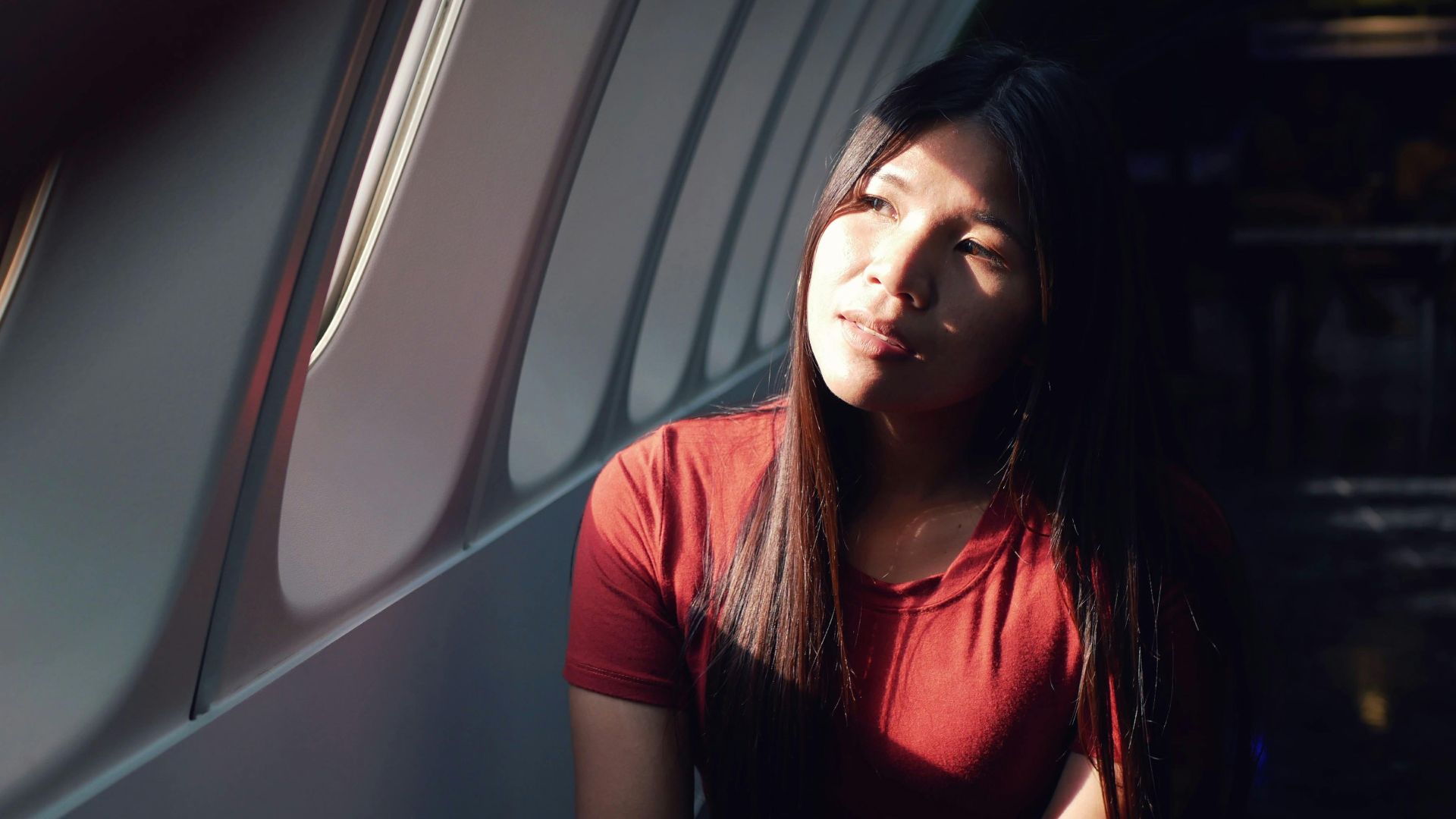 A woman in a red shirt sits in an airplane, gazing thoughtfully out the window as sunlight illuminates her face.