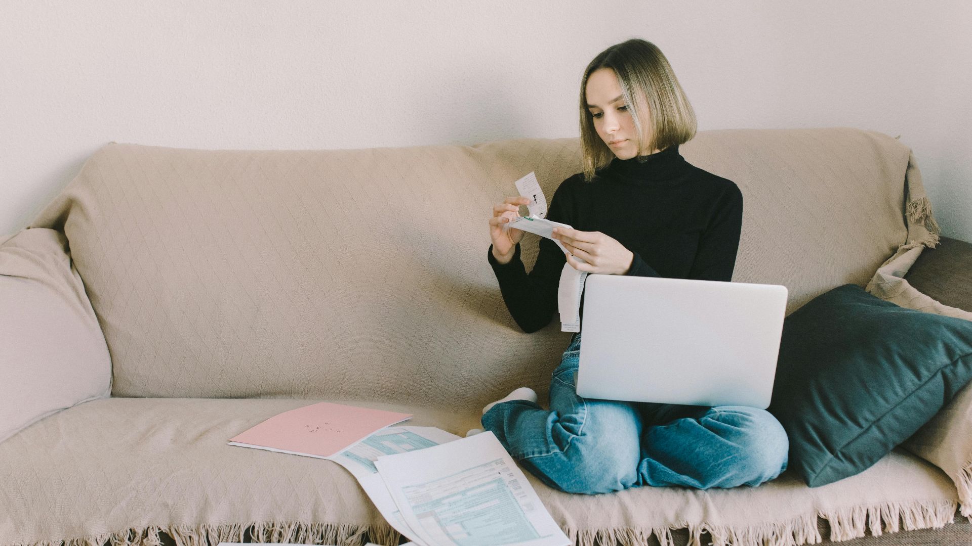 A woman working from home on a sofa with a laptop and documents, managing finances.