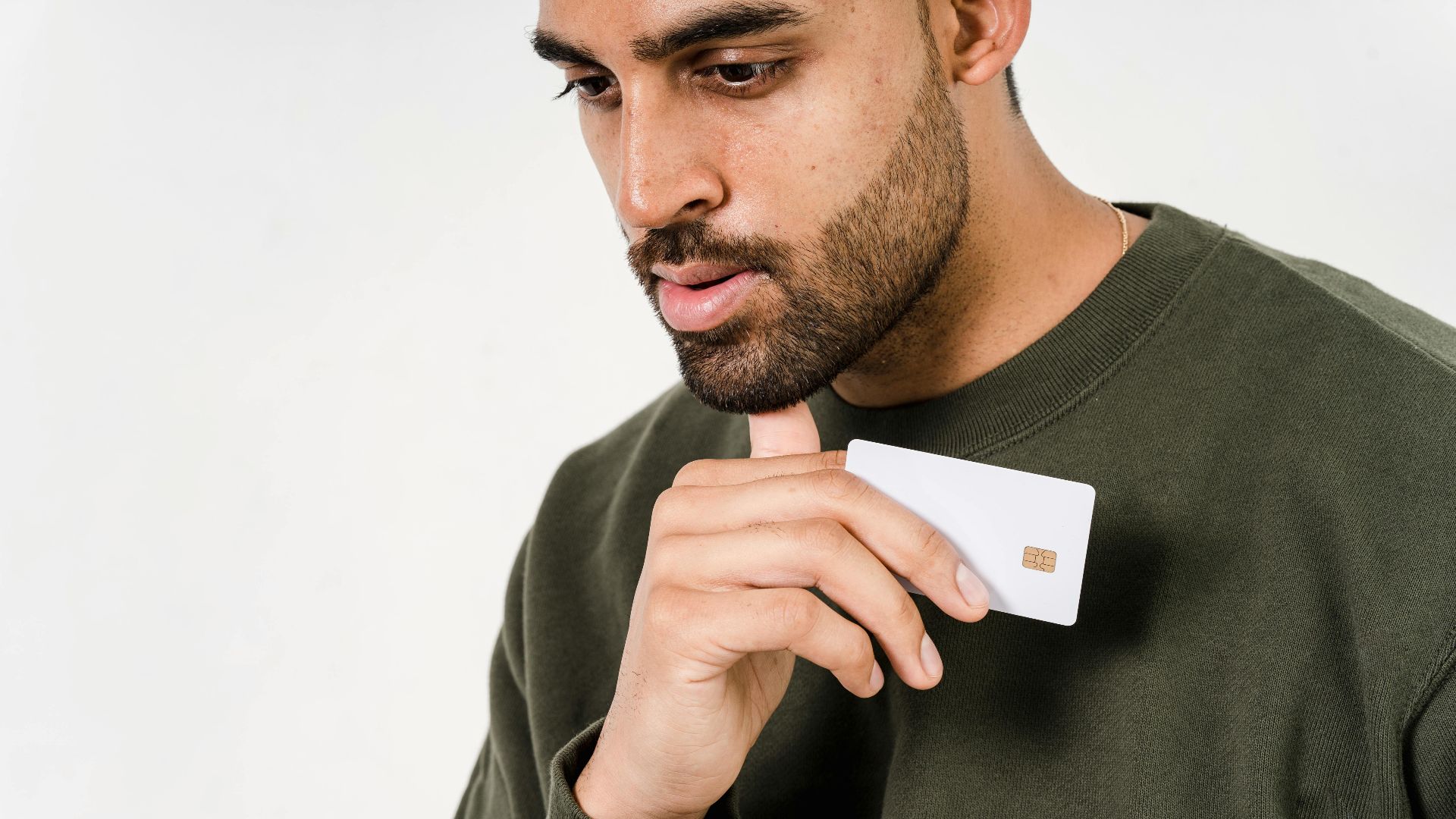 Pensive man in green sweater holds a white credit card against a white background.