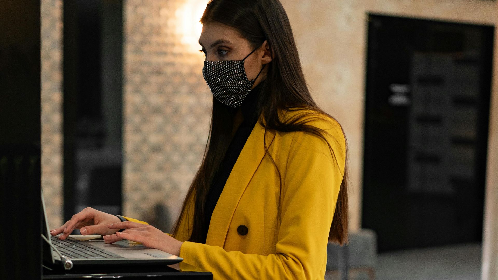 Woman wearing mask and yellow coat using laptop in hotel lobby next to suitcase.