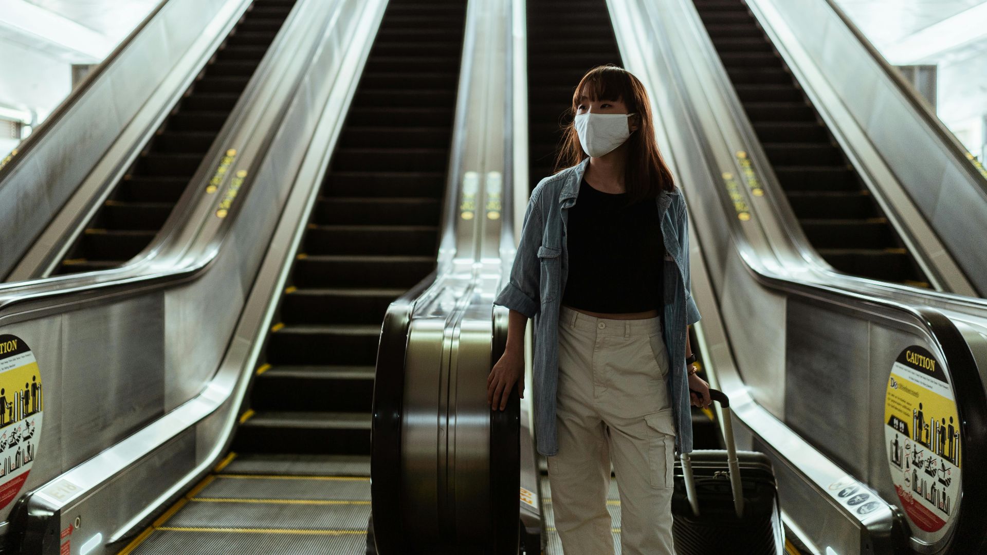Asian woman wearing a face mask while traveling on an airport escalator during the pandemic, carrying luggage.