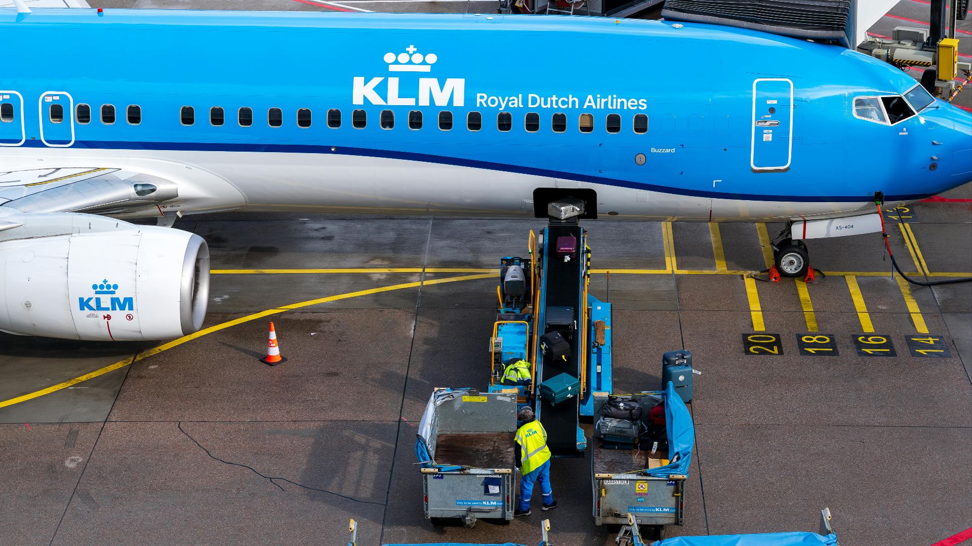 KLM airplane being loaded at a terminal gate with luggage carts and workers preparing for flight.