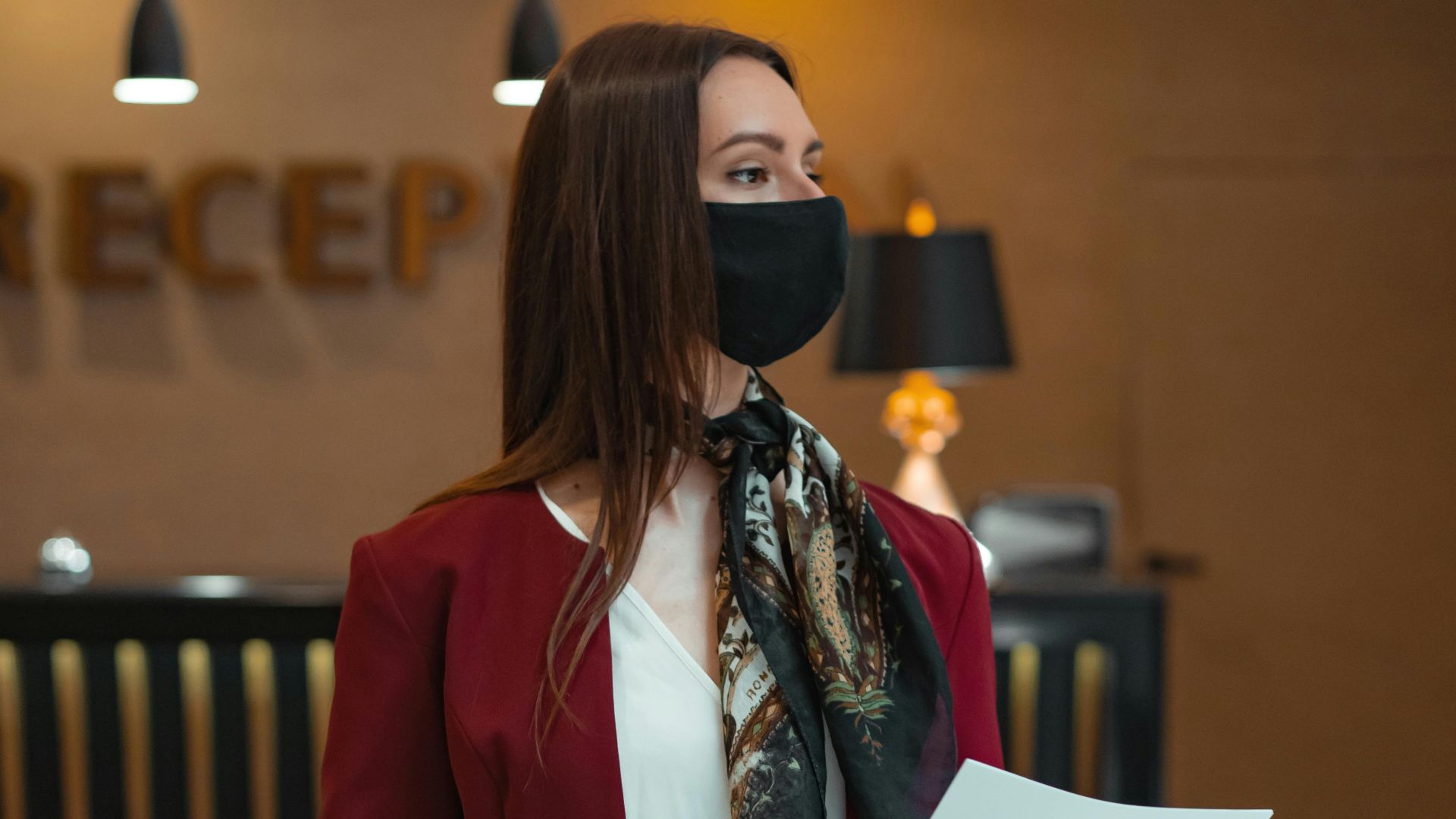 Woman wearing a mask and holding papers at hotel reception, conveying safety and preparedness.