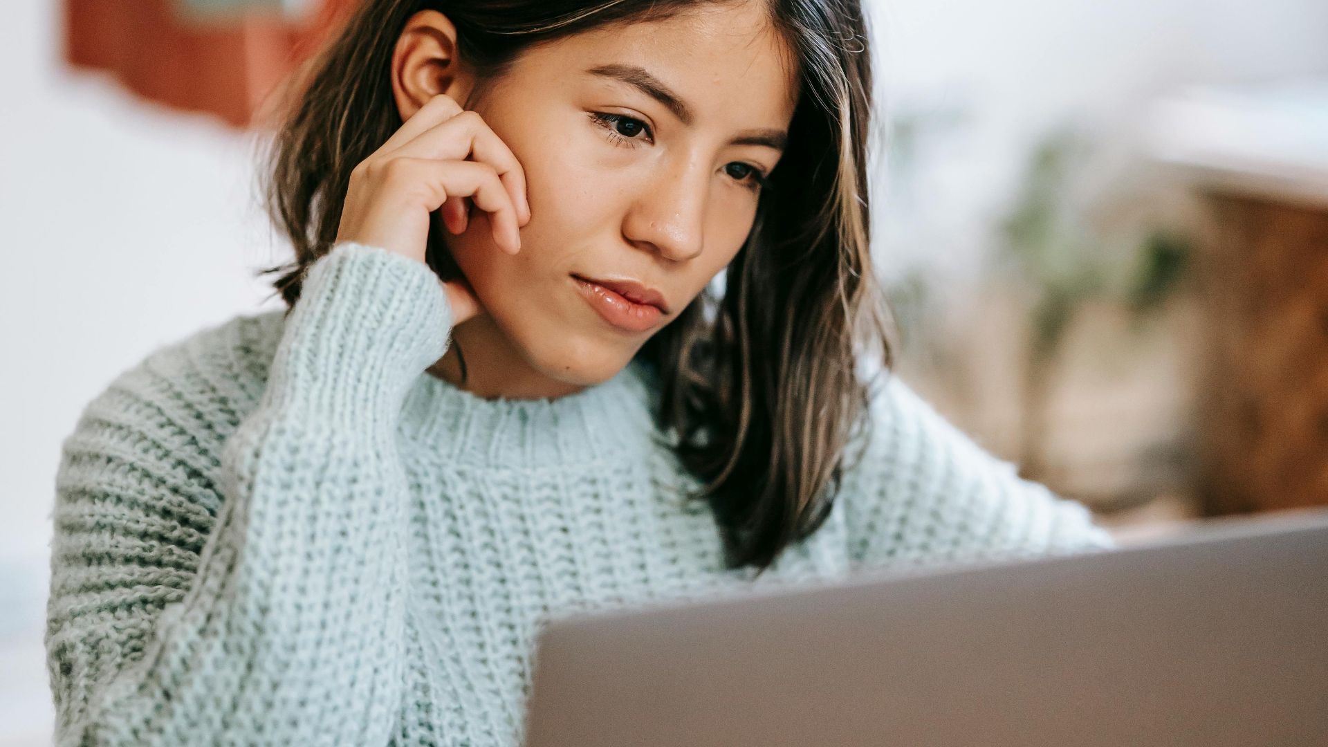 Youthful Latin American female in casual clothes sitting at table with laptop while looking at screen attentively at home