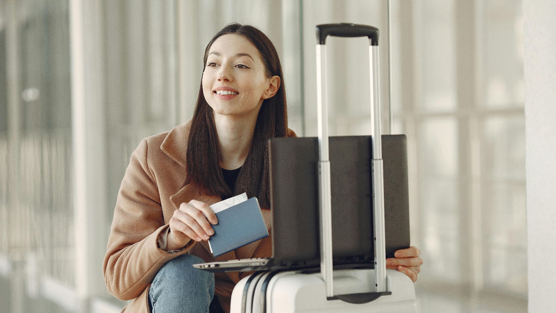 A woman sitting with her laptop and luggage, holding a passport, waiting in an airport terminal.
