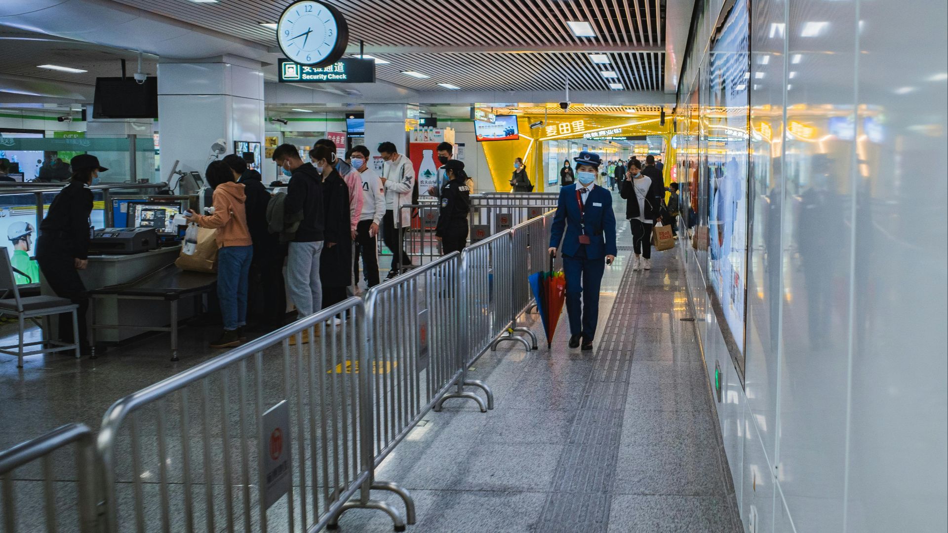 Travelers passing through airport security with staff and metal railings visible.