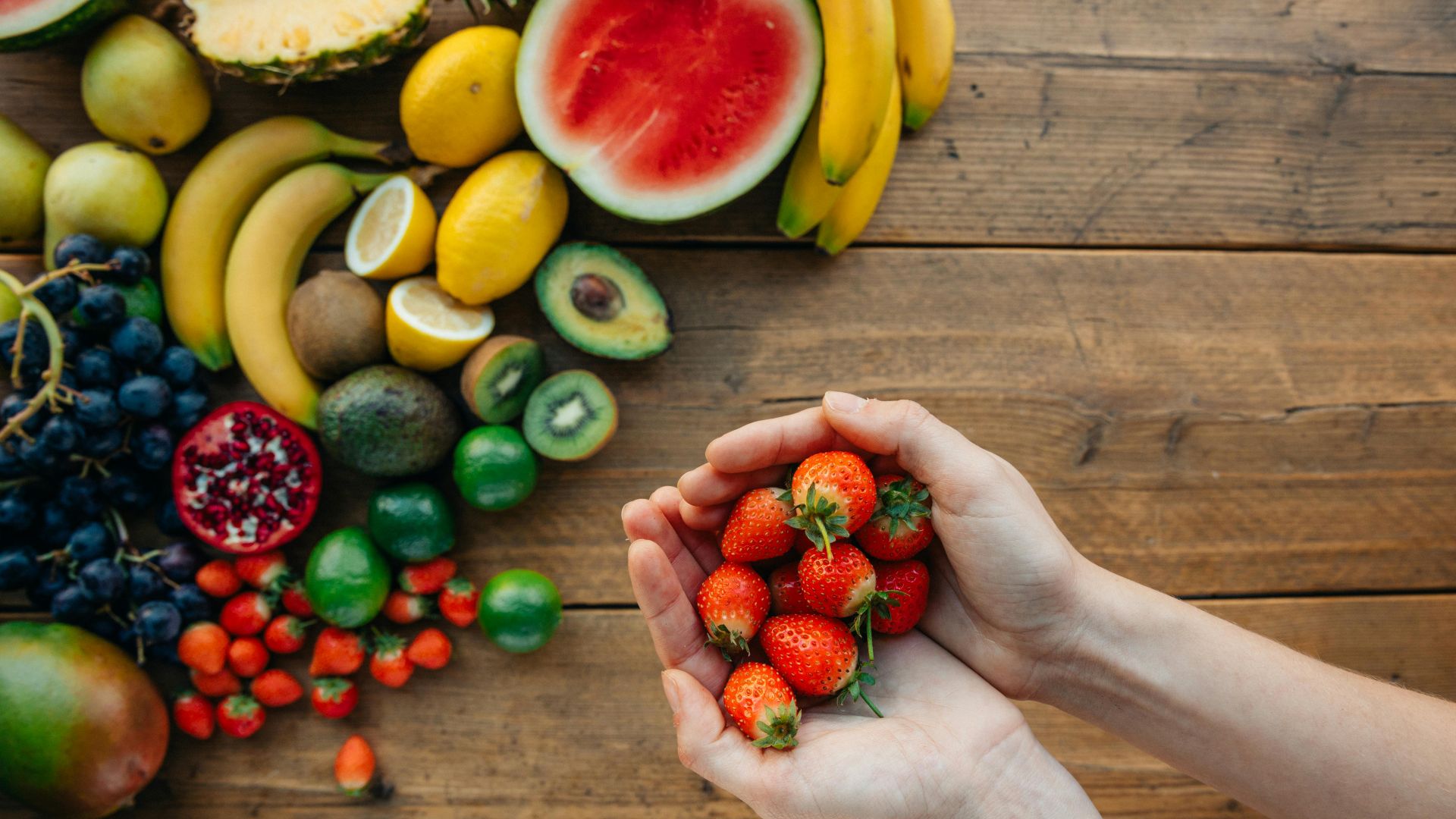 Close-up of hands holding fresh strawberries with assorted colorful fruits on a wooden table.