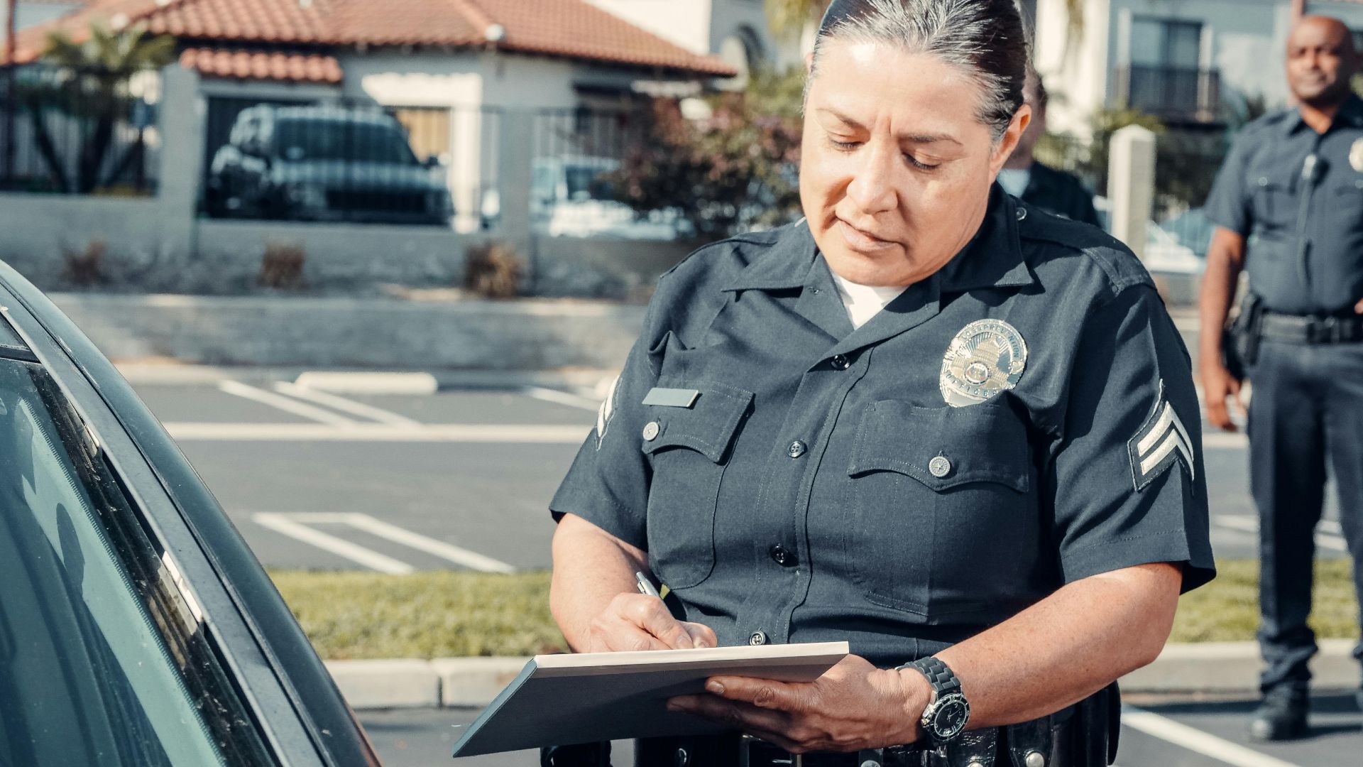 Police officer writing a ticket to a car in a sunny parking lot with a colleague in the background.