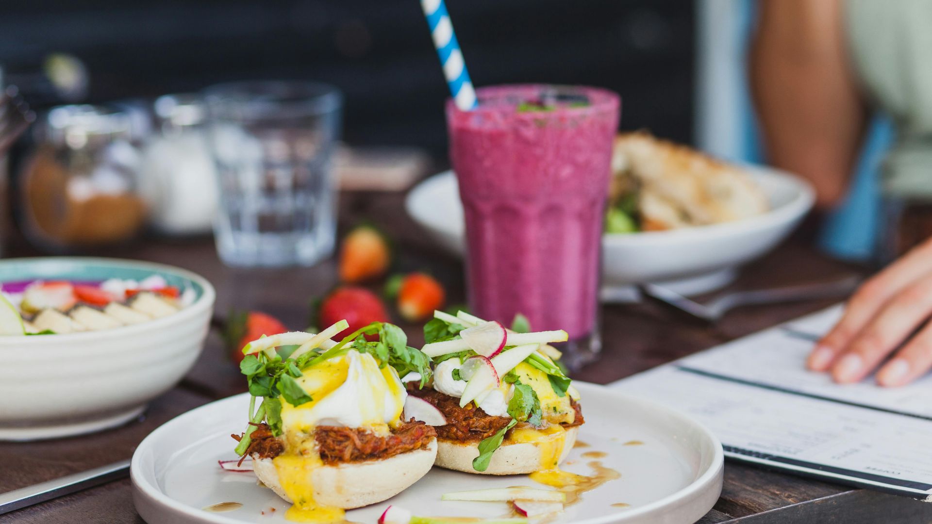 Appetizing sandwiches with poached eggs and sauce served with berry smoothie and placed on table with crop unrecognizable woman reading menu on background