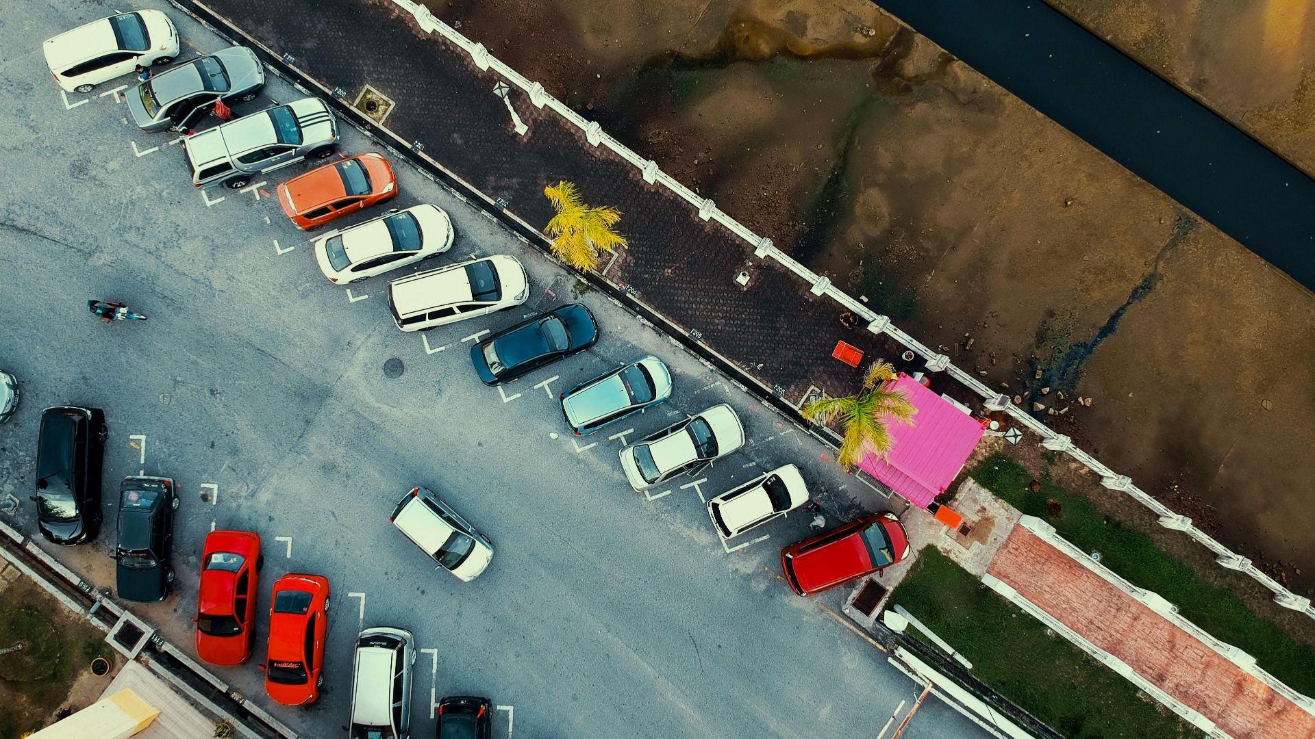 Aerial shot of a parking lot with cars and a pink building, next to a waterway.